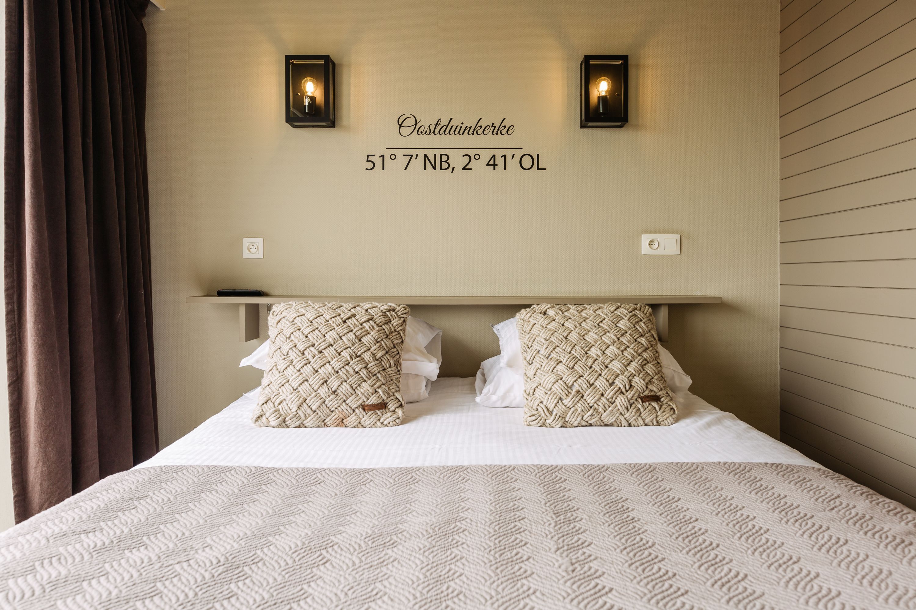 A neatly made bed with a patterned bedspread and two decorative pillows, a wall-mounted light fixture, and a sign with coordinates.
