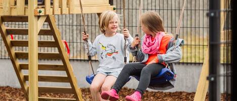 A wooden play structure with swings and a slide, surrounded by mulch and grass.