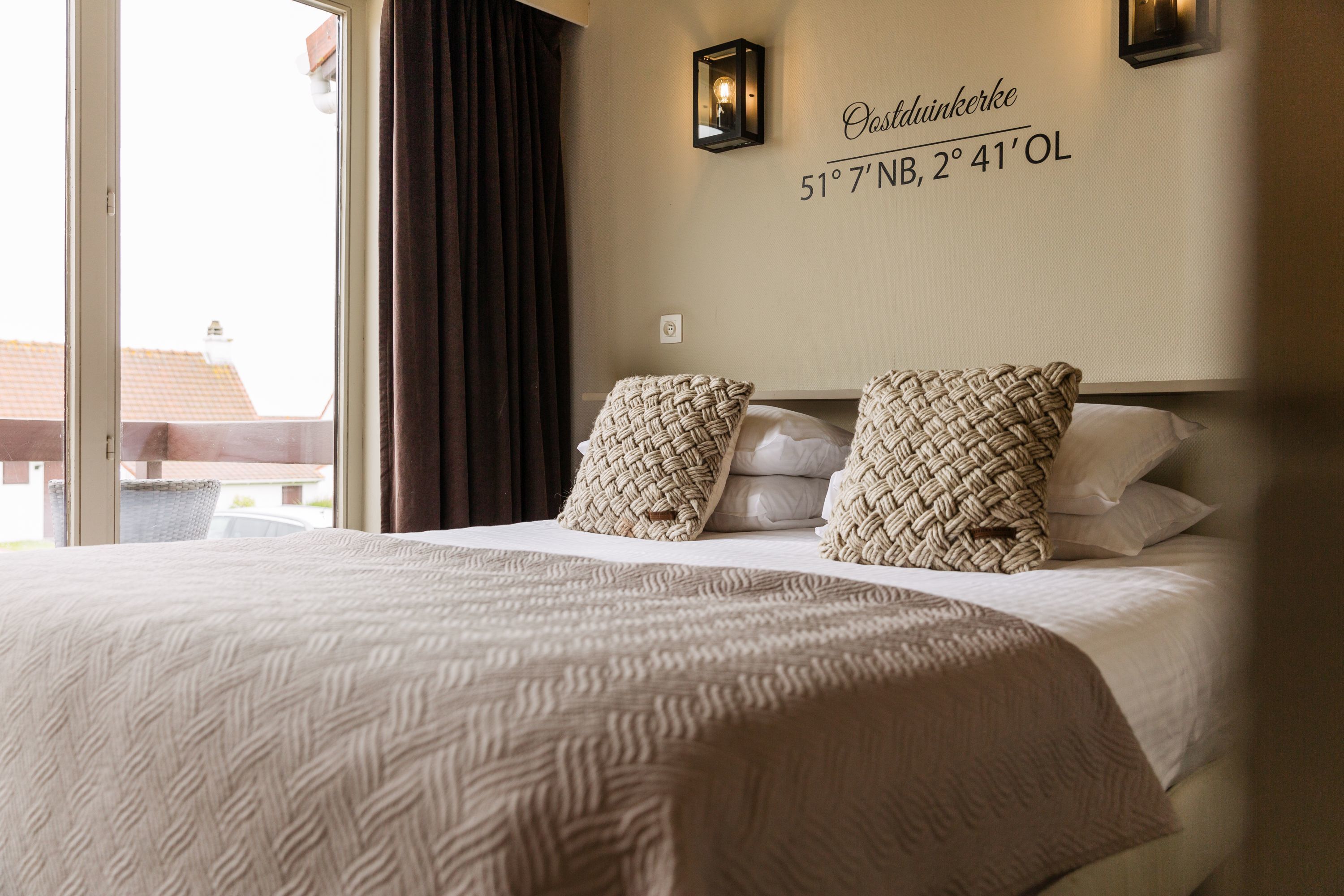 Habitación de hotel con una cama, dos almohadas tejidas, una lámpara de pared y un cartel que indica la ubicación.