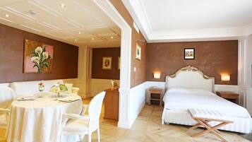A hotel room with a round dining table, a bed with an ornate headboard, and a small bedside table.
