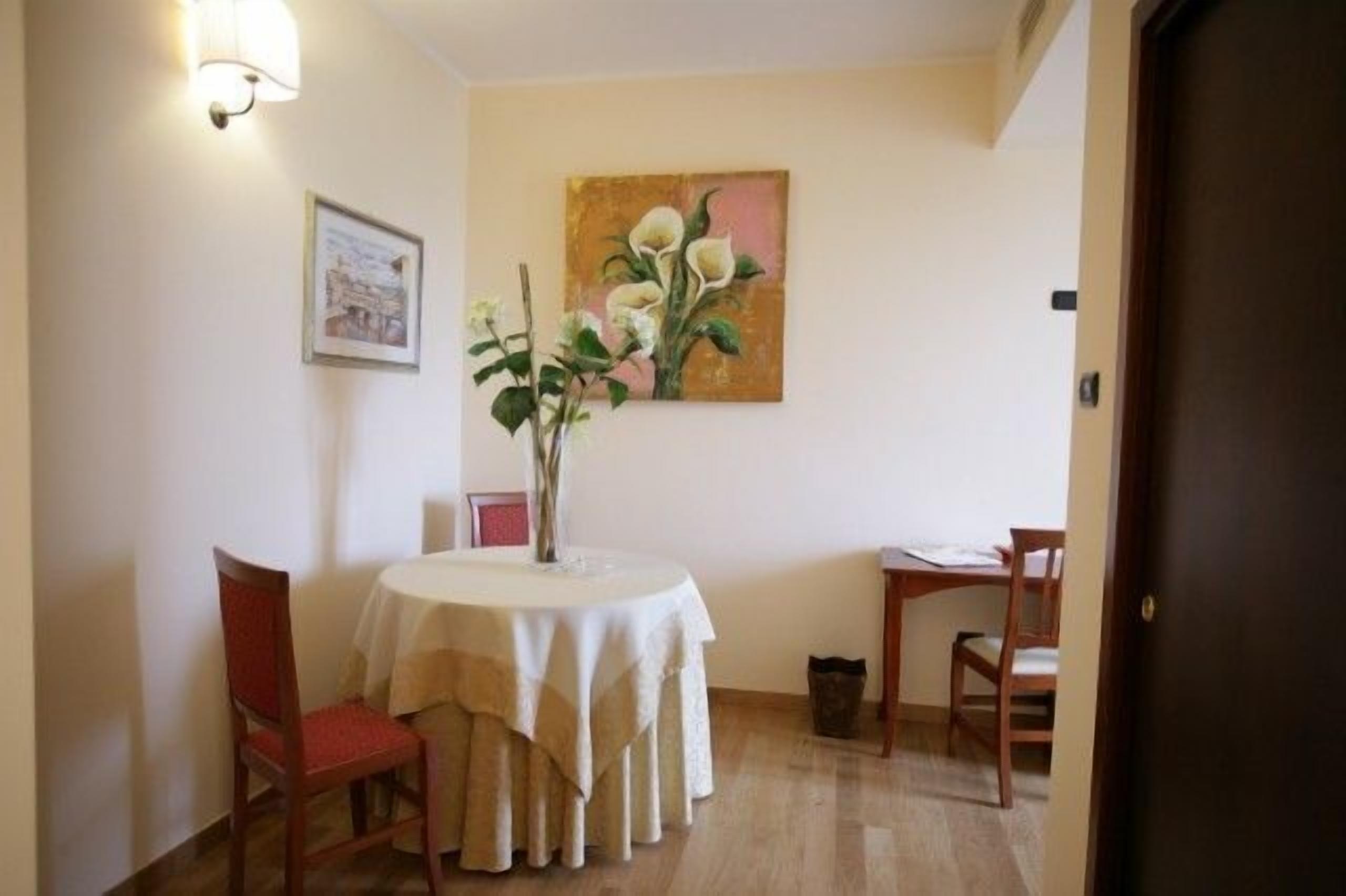 A dining area with a round table covered with a white tablecloth, two chairs, a vase with flowers, and a painting on the wall.