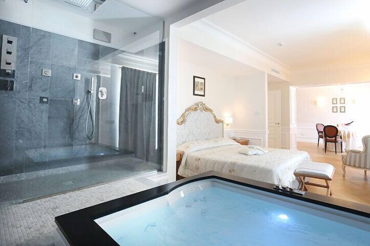 Una habitación de hotel moderna con ducha de vidrio, una cama grande con cabecera blanca y un área de descanso con mesa y sillas.