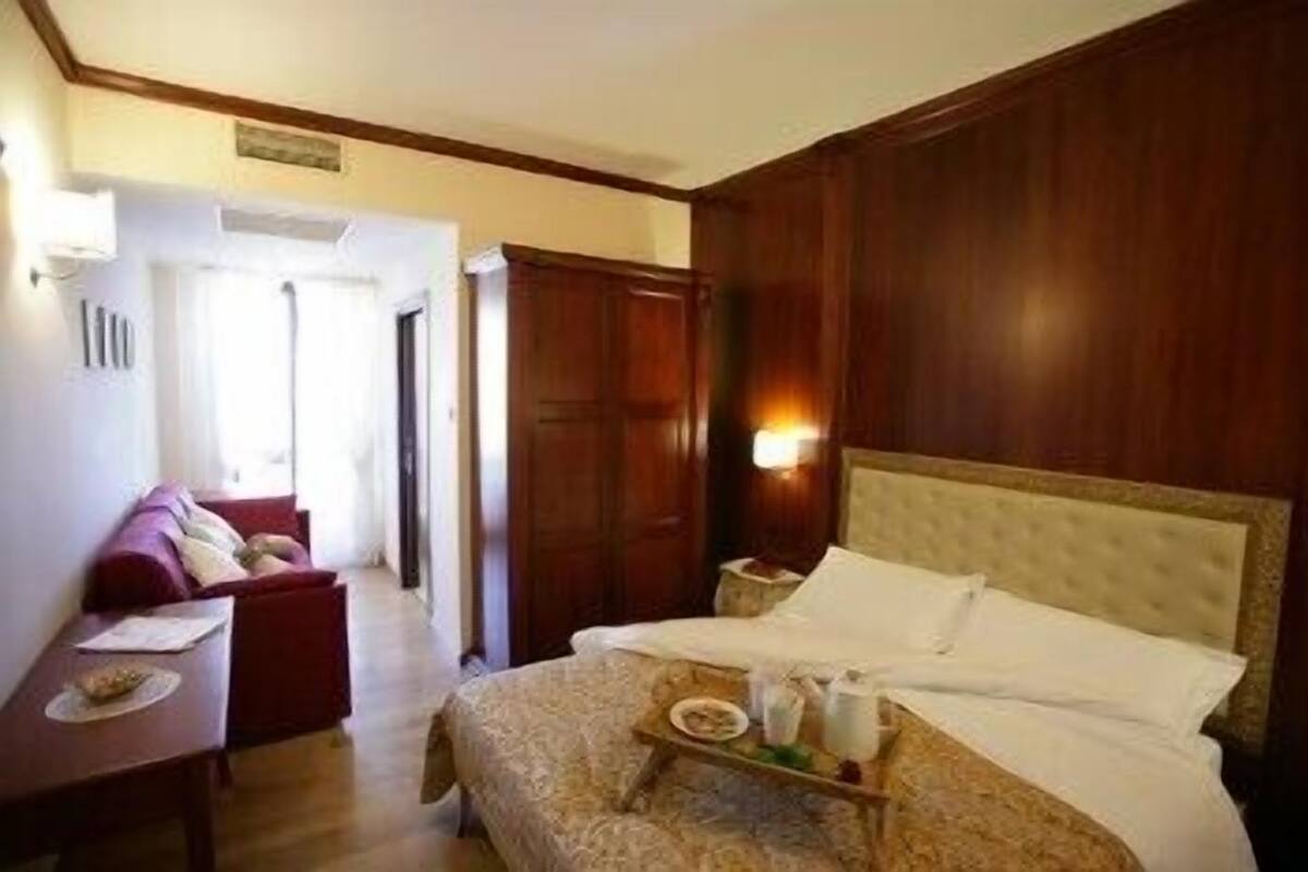 Habitación de hotel con una cama grande, una mesita de noche con un servicio de desayuno, un armario de madera y una zona de estar con sofá y mesa.