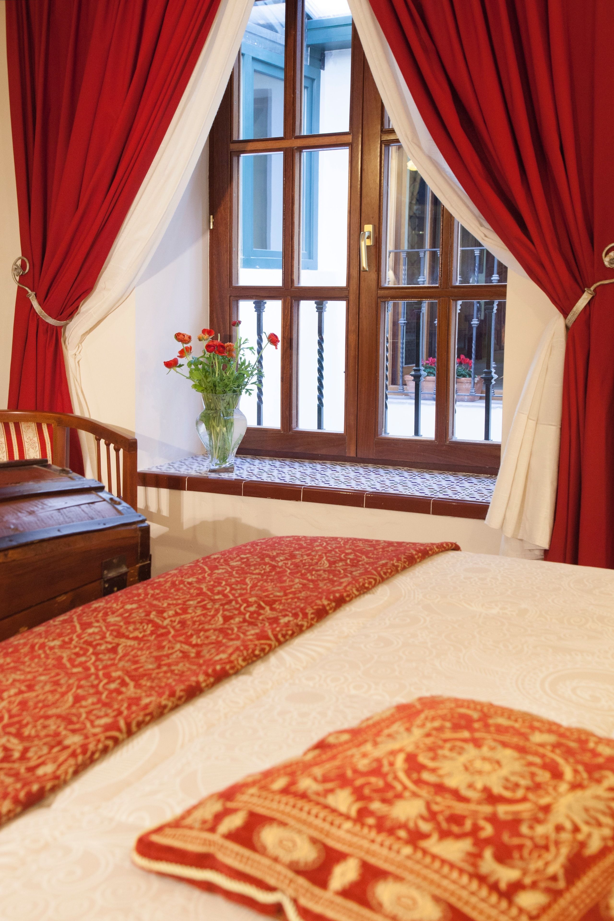 Habitación de hotel con cama, mesita de noche de madera, un jarrón con flores y una ventana con cortinas rojas.
