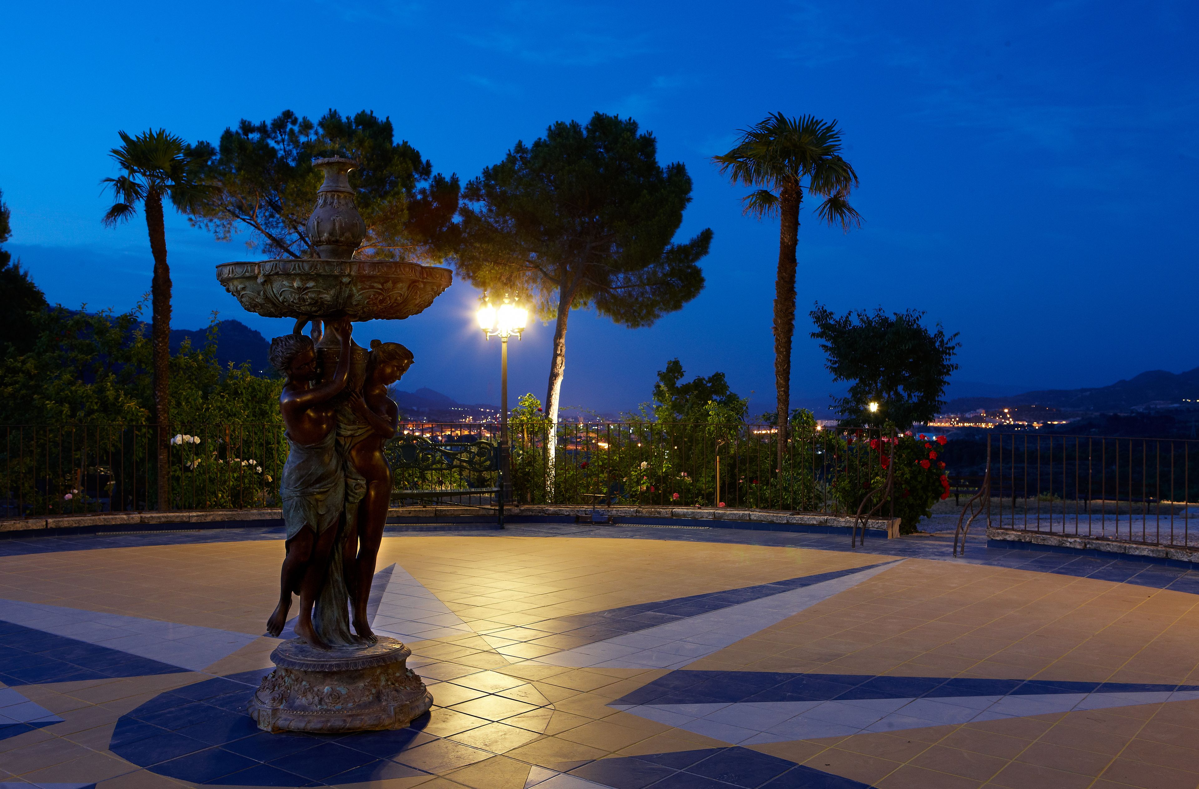Un espacio al aire libre iluminado con una fuente, suelo de baldosas y vista a un paisaje urbano de fondo.