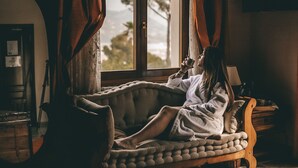 Una persona descansando en un banco acolchado junto a una ventana con cortinas, una lámpara y una mesa de madera con una cesta.