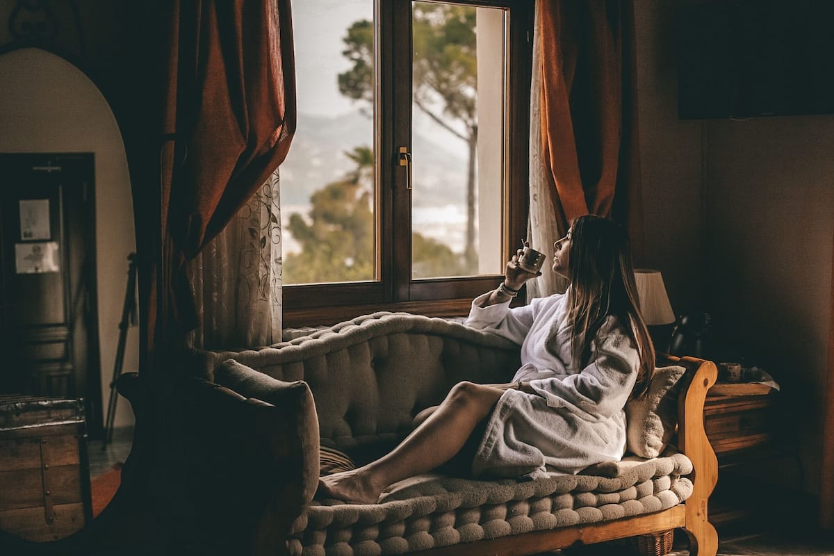 A person relaxing on a cushioned bench by a window with curtains, a lamp, and a wooden table with a basket.
