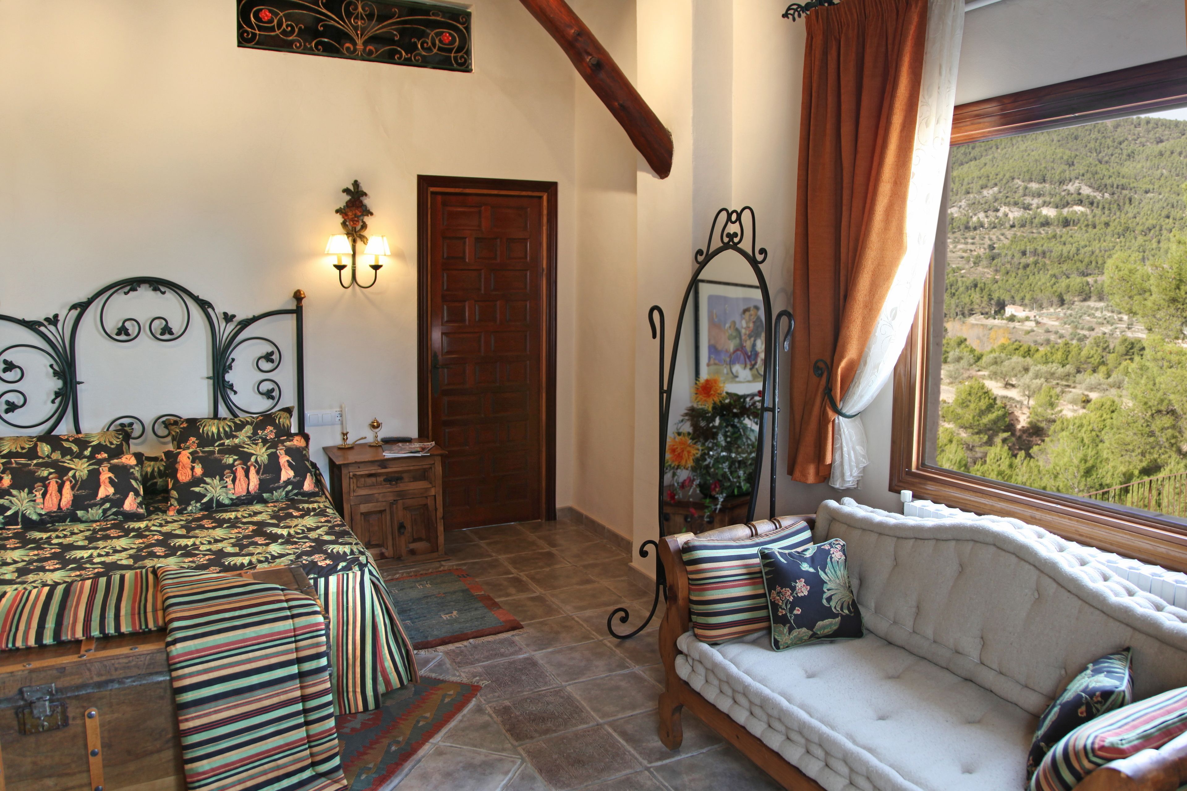 A bedroom with a bed, a sofa, a wooden door, a mirror, and a view of a mountainous landscape.
