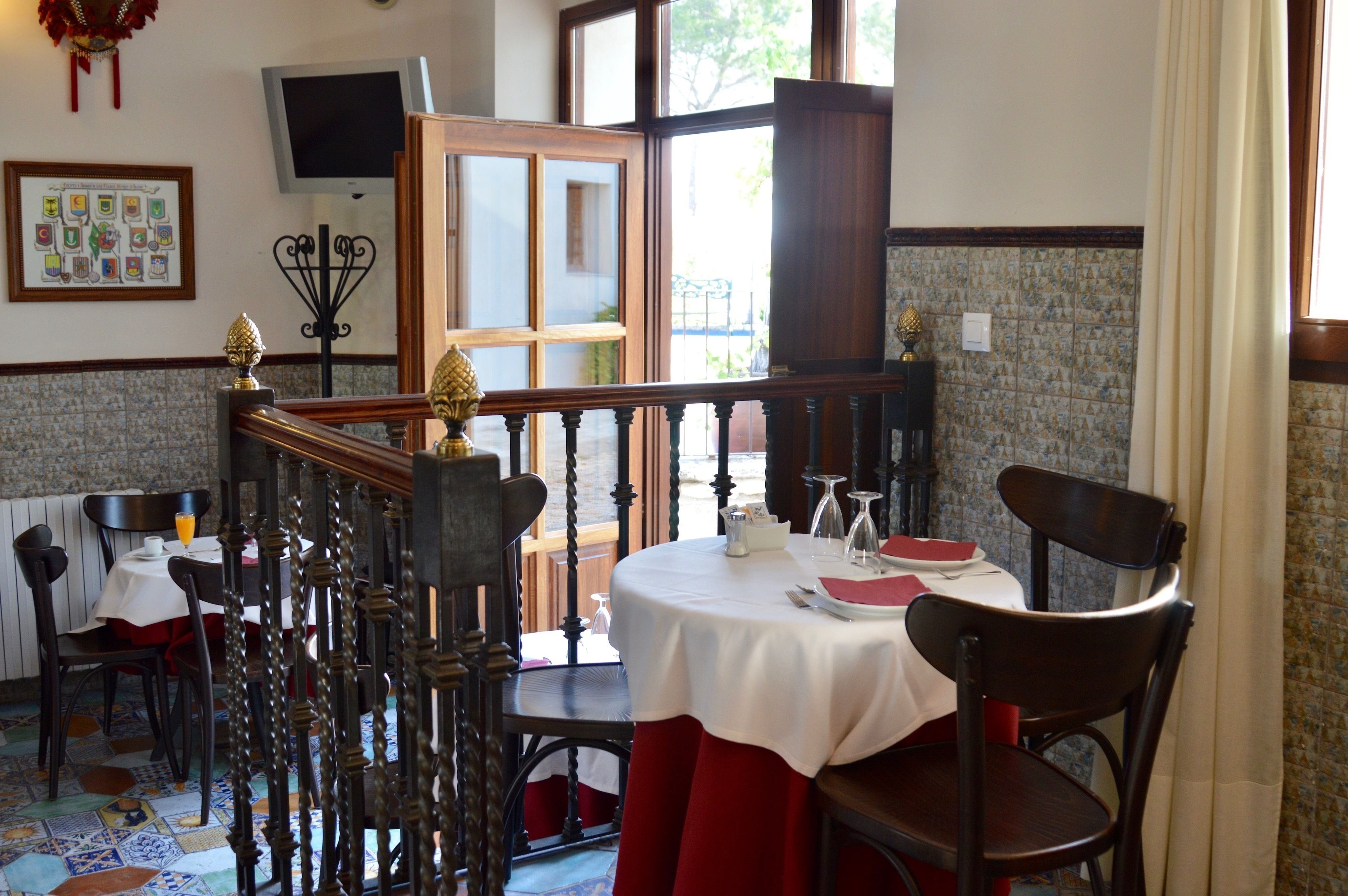 A dining area with a round table set for two, wooden chairs, a television on the wall, and a window with curtains.
