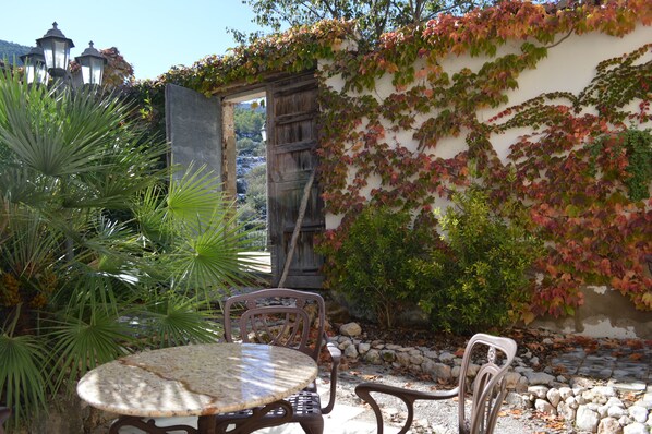 Un patio con una mesa redonda y dos sillas, rodeado de abundante vegetación y un muro de piedra.