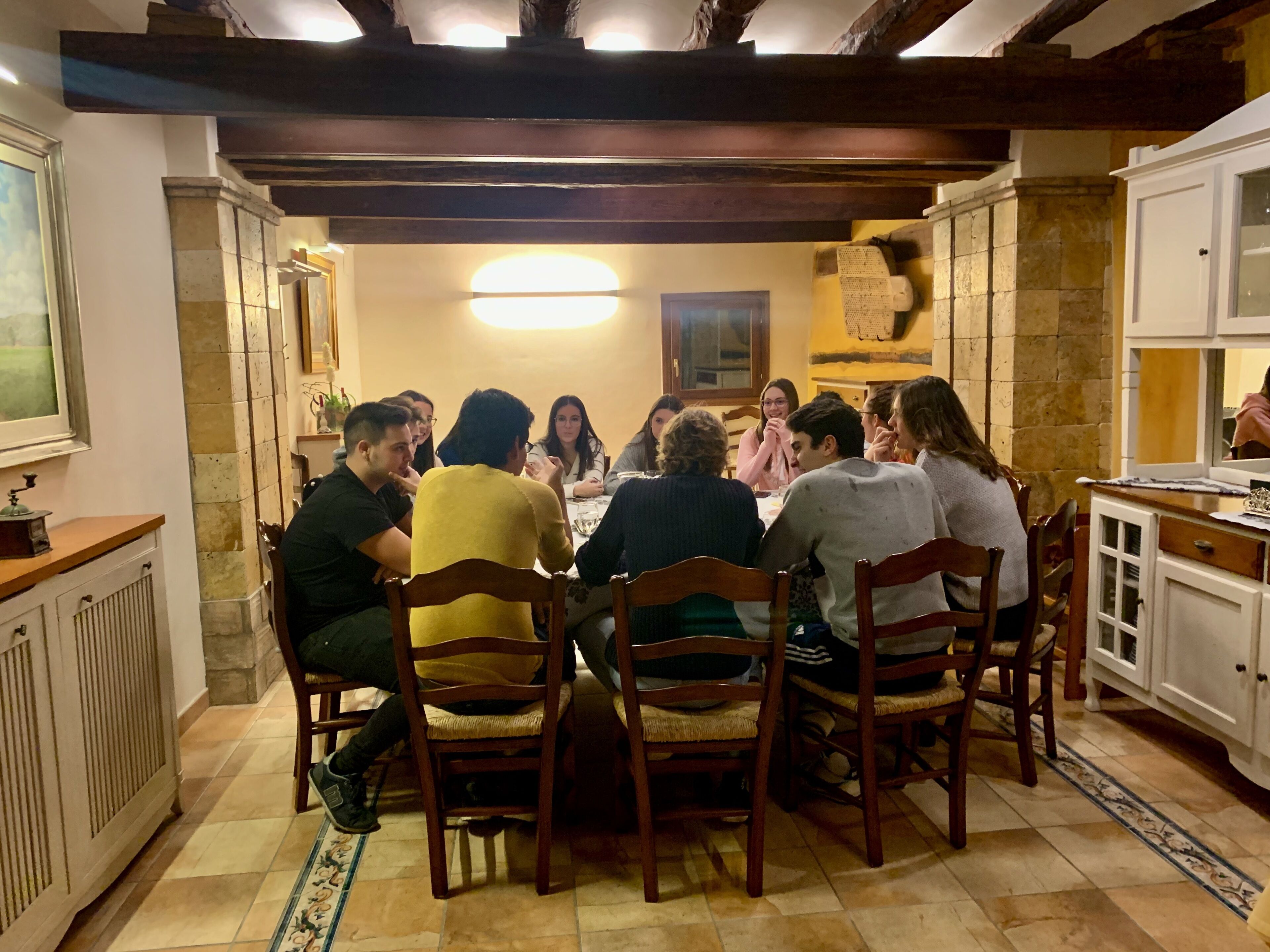 A group of people seated around a table in a room with stone walls, wooden beams, and a tiled floor.