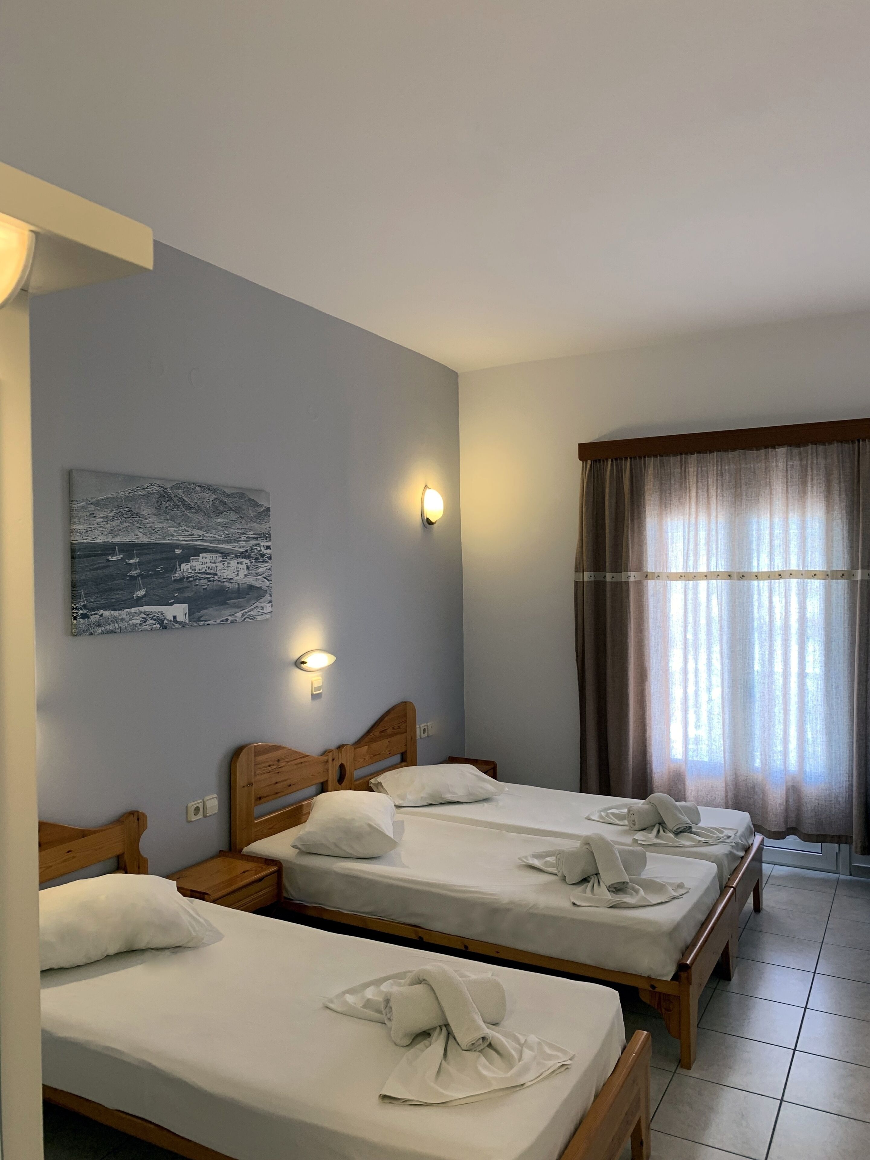 A hotel room with three wooden beds, a painting on the wall, and a window with curtains.
