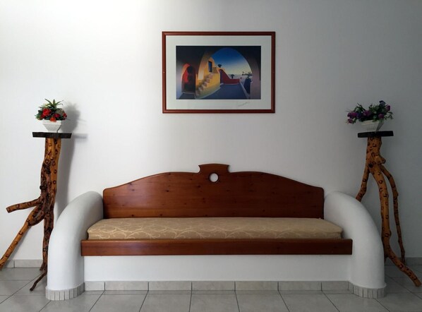 A white sofa with a wooden headboard, flanked by two wooden stands with flower pots. A framed painting hangs above the sofa.