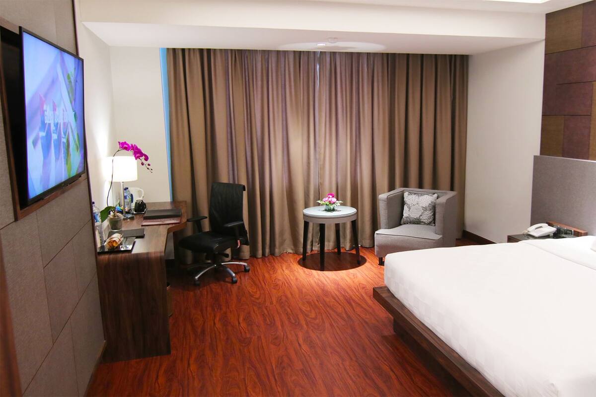 A hotel room with a bed, a desk with a chair, a TV, a small table with a flower, and a curtain.