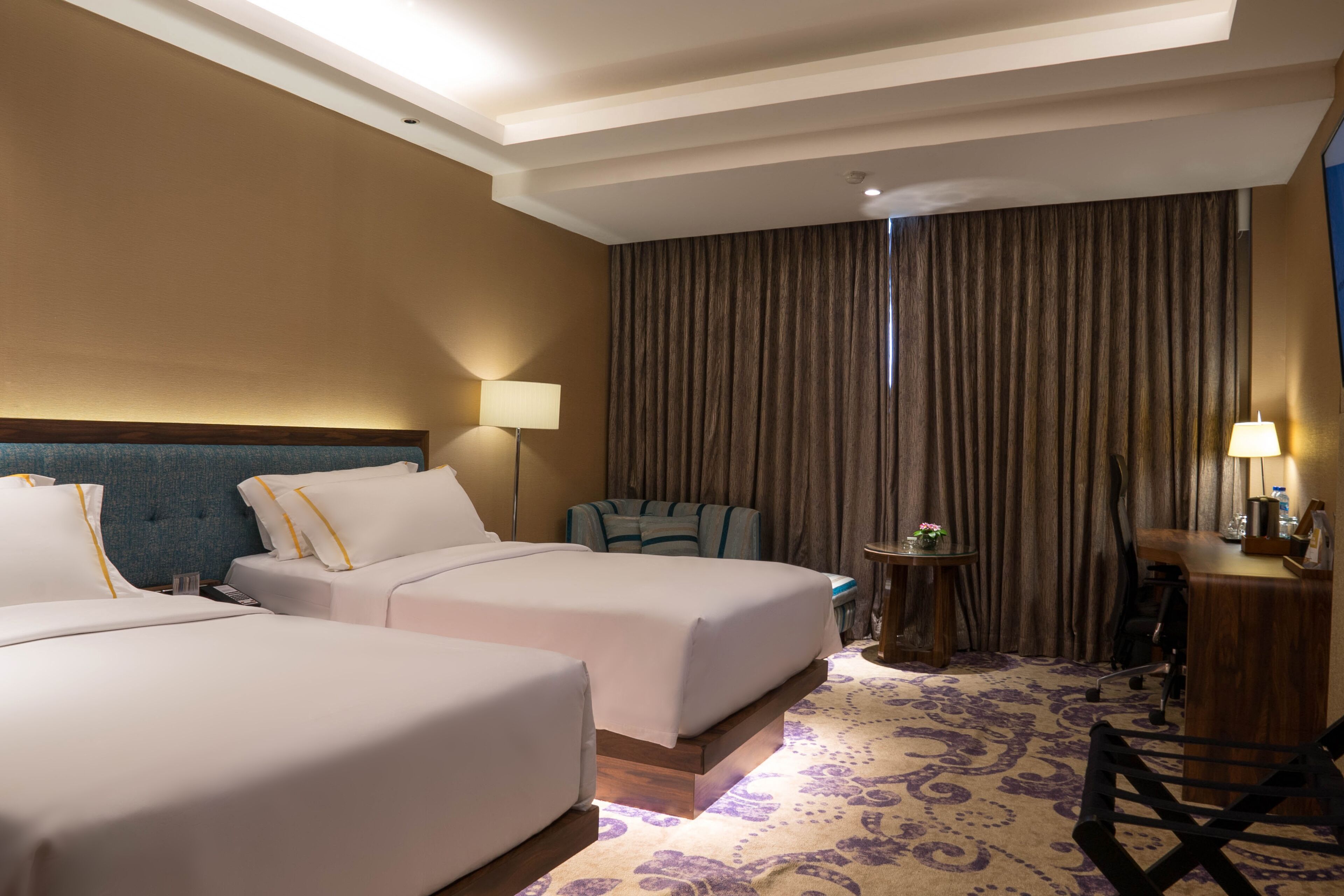 Ein Hotelzimmer mit zwei Betten, einem Schreibtisch, einem Stuhl, einem kleinen Tisch und einem großen Fenster mit Vorhängen.