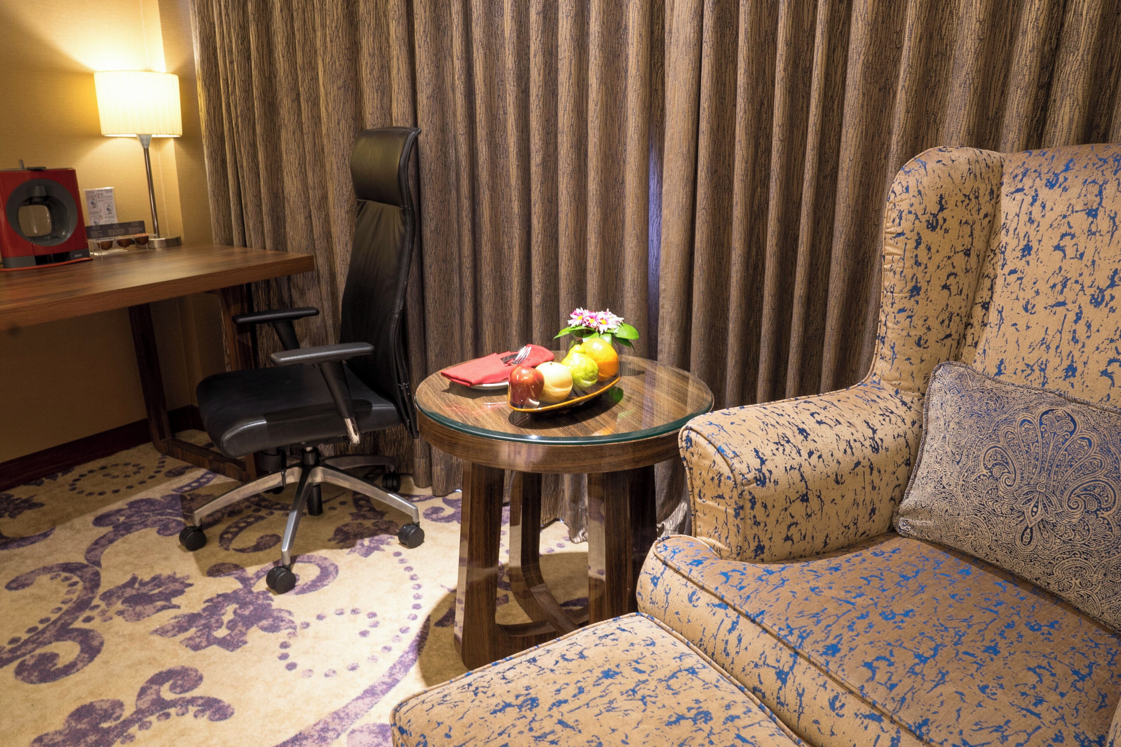 A hotel room with a sofa, a small round table with a fruit bowl, a chair, a desk with a lamp, and a curtain.