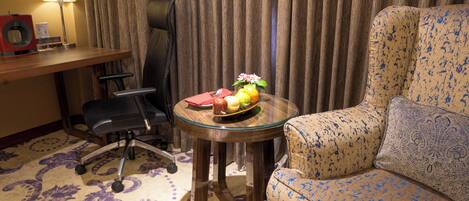 A hotel room with a sofa, a small round table with a fruit bowl, a chair, a desk with a lamp, and a curtain.