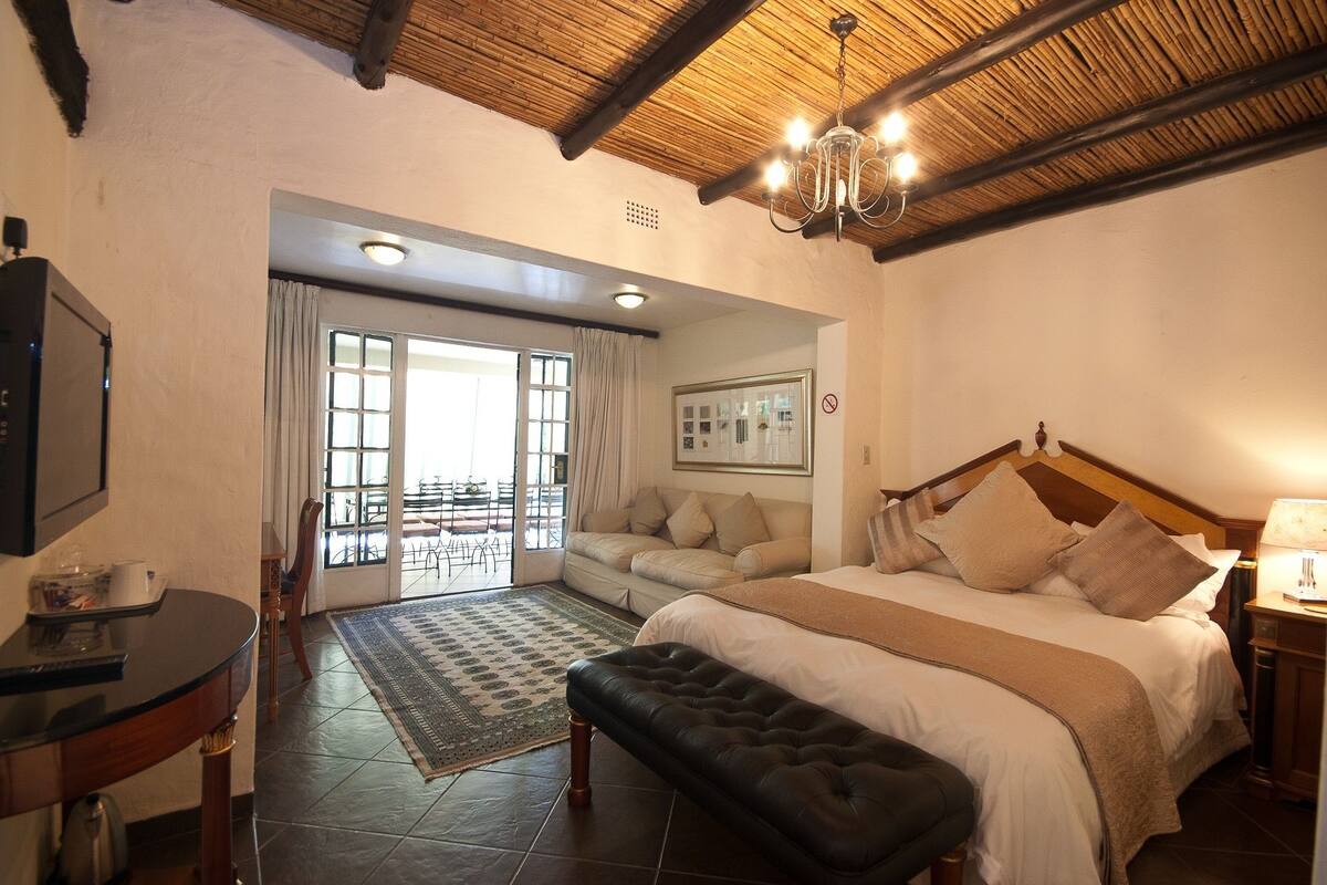 A bedroom with a large bed, a wooden ceiling, a chandelier, a TV, and a view through a glass door to an outdoor area.