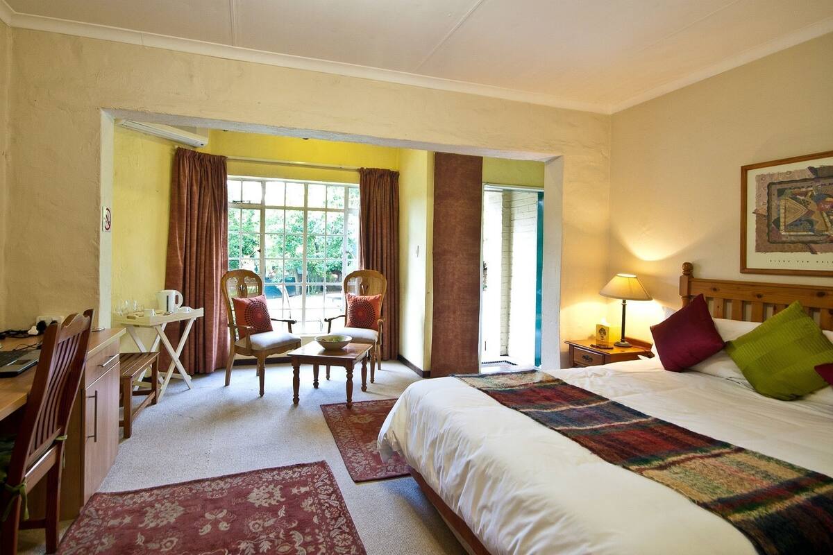 A hotel room with a bed, a desk with a computer, a chair, a table with a bowl, and a view of the outdoors through a door.