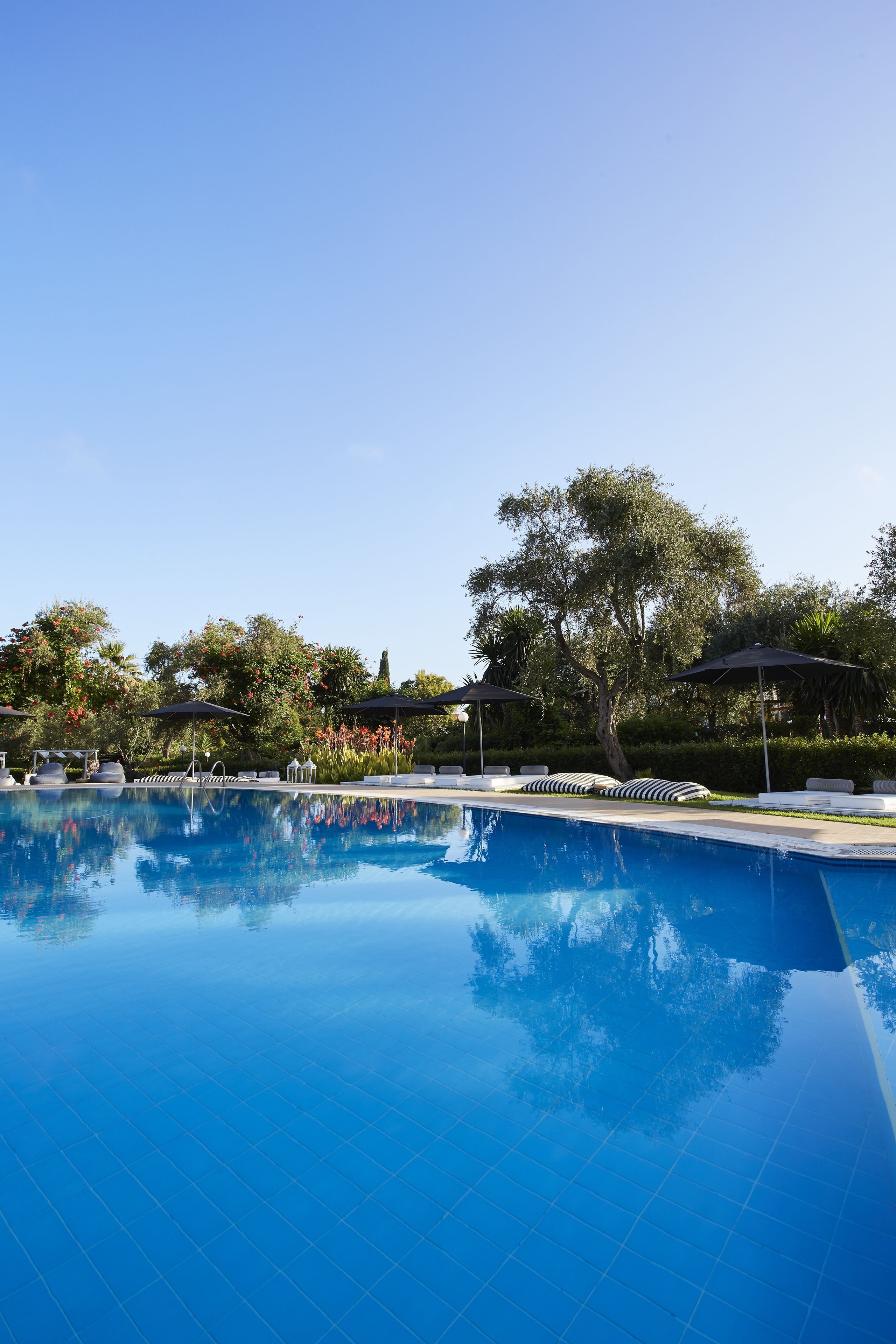 Piscina azul e límpida com espreguiçadeiras e guarda-sóis, rodeada por árvores e vegetação.