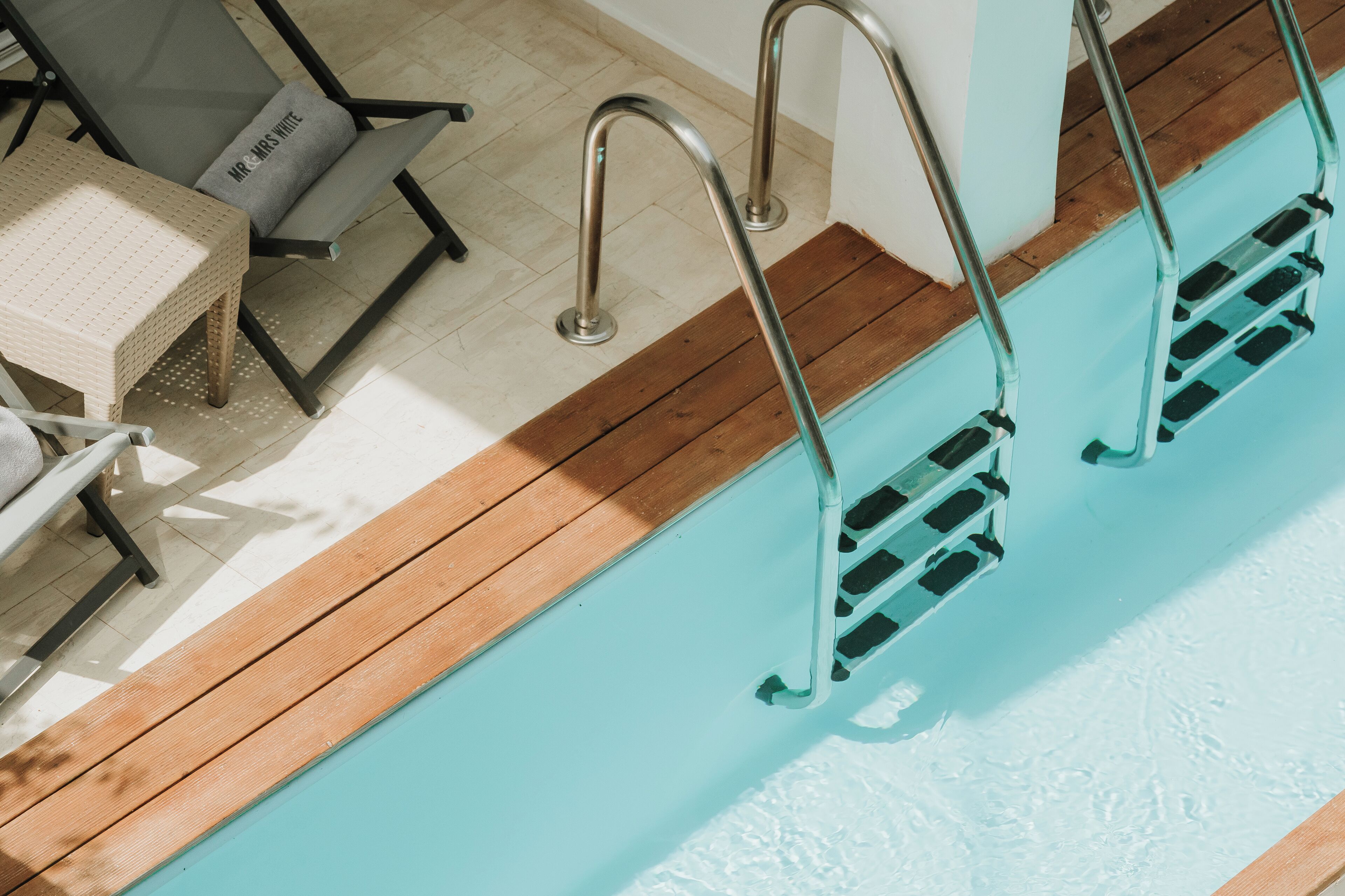 Un espace aménagé au bord de la piscine, avec une terrasse en bois, un escalier métallique pour accéder à la piscine et une chaise longue dotée d’un coussin.