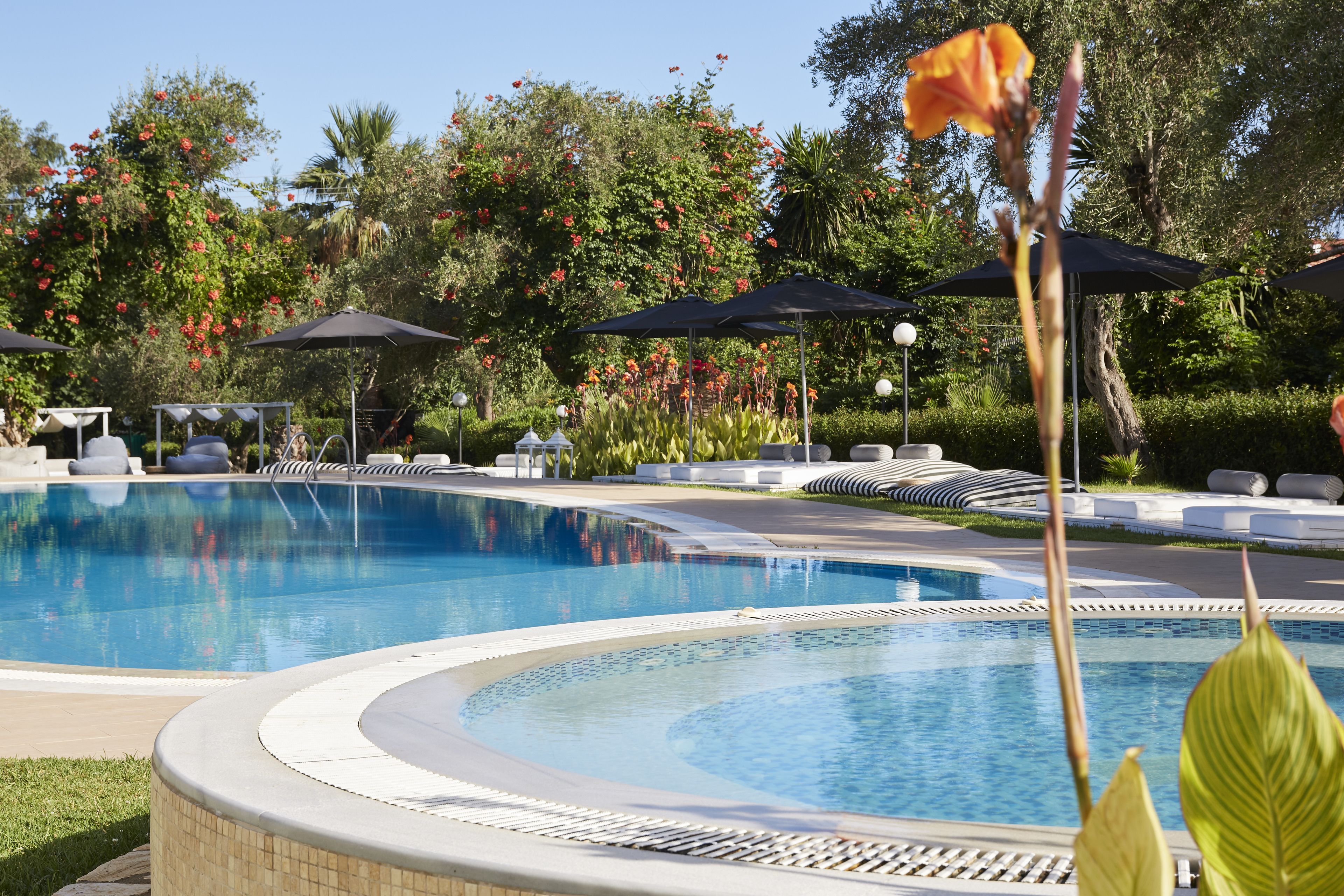 Piscina con sillas de descanso y sombrillas, rodeada de exuberante vegetación y árboles.