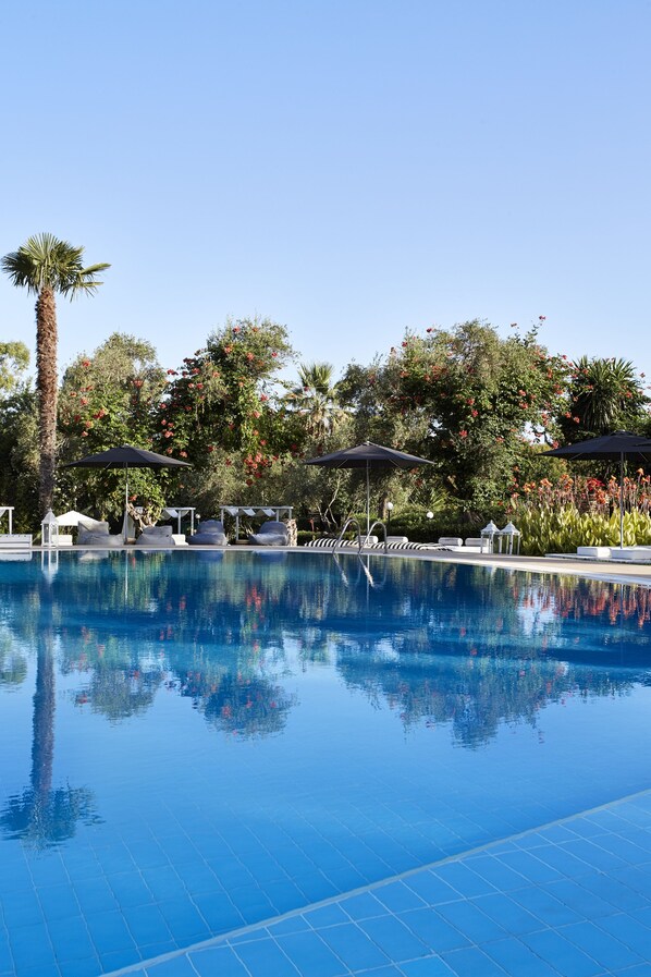 A clear blue pool with lounge chairs and umbrellas, surrounded by lush greenery and trees.