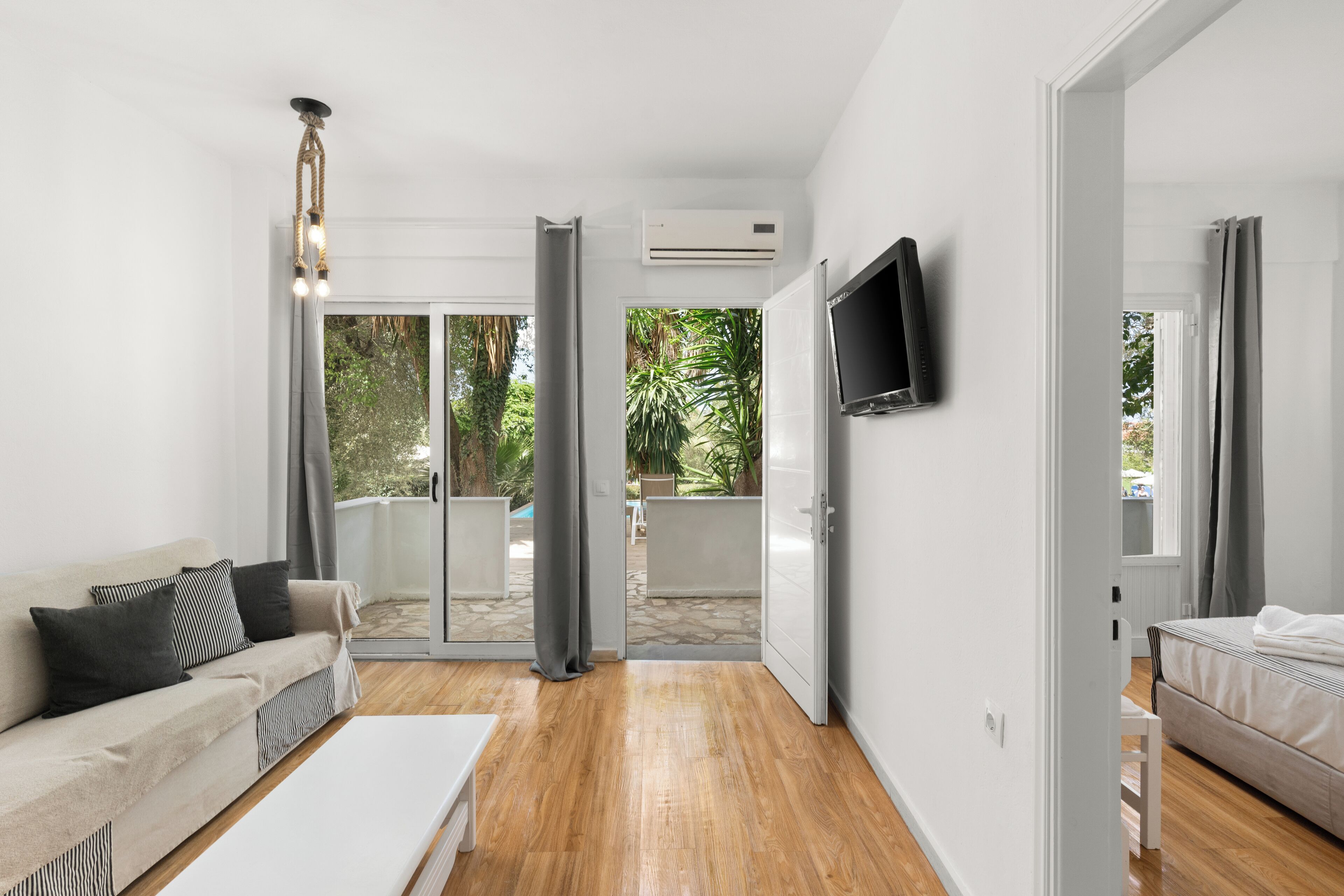 Una sala de estar moderna con un sofá, una mesa de centro y un televisor montado en la pared. Hay un ventanal corredero que da a un espacio exterior con piscina y vegetación.