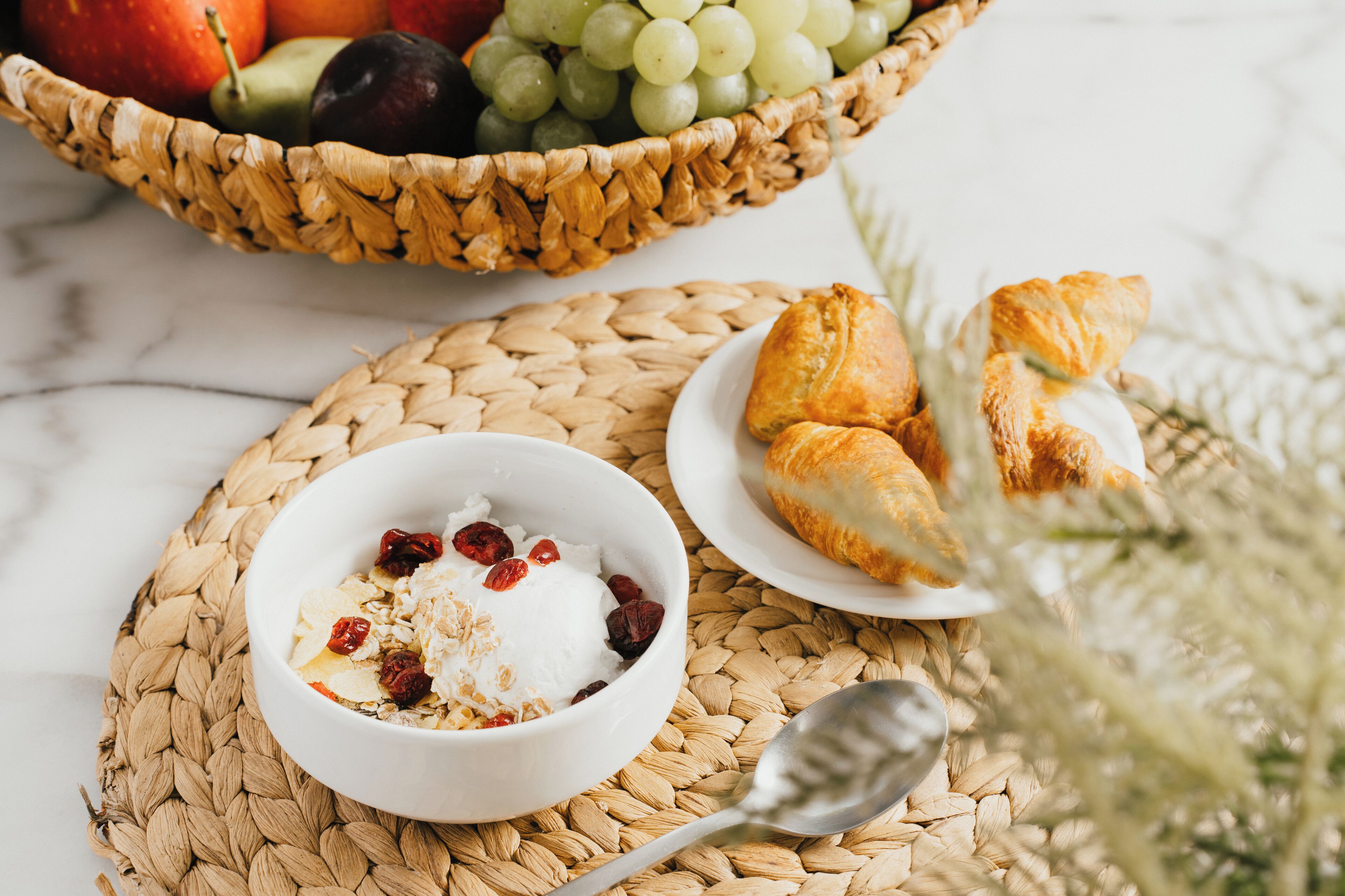 Ein Frühstückstisch mit gewebter Unterlage, einer Schüssel mit Frühstücksflocken und Trockenfrüchten, einer Auswahl an Croissants und einem Obstkorb.