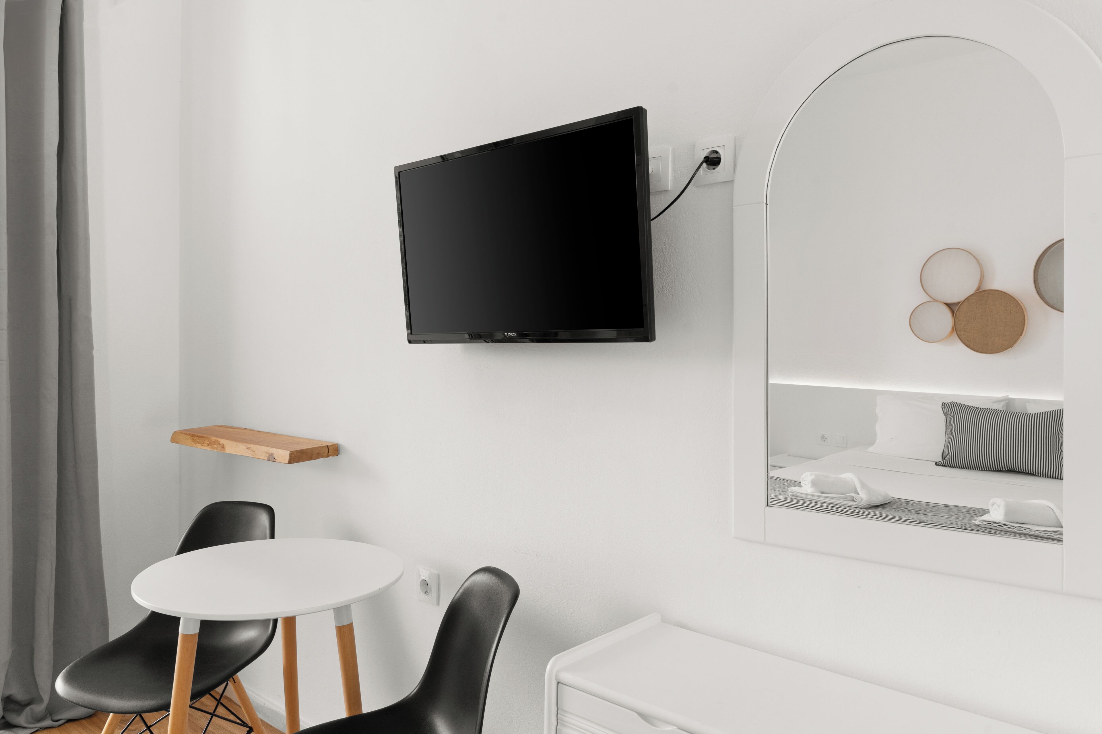 Ett modernt hotellrum med en platt-TV som är monterad på väggen, en liten hylla i trä, ett runt vitt bord med två svarta stolar och en säng med en spegel som reflekterar rummet.