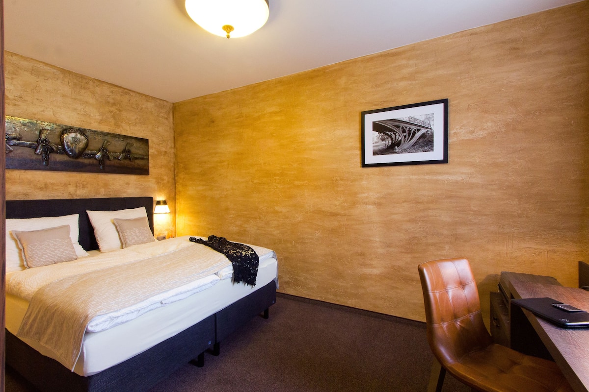 A hotel room with a bed, a desk, and a chair. There is a framed picture on the wall and a decorative piece above the bed.
