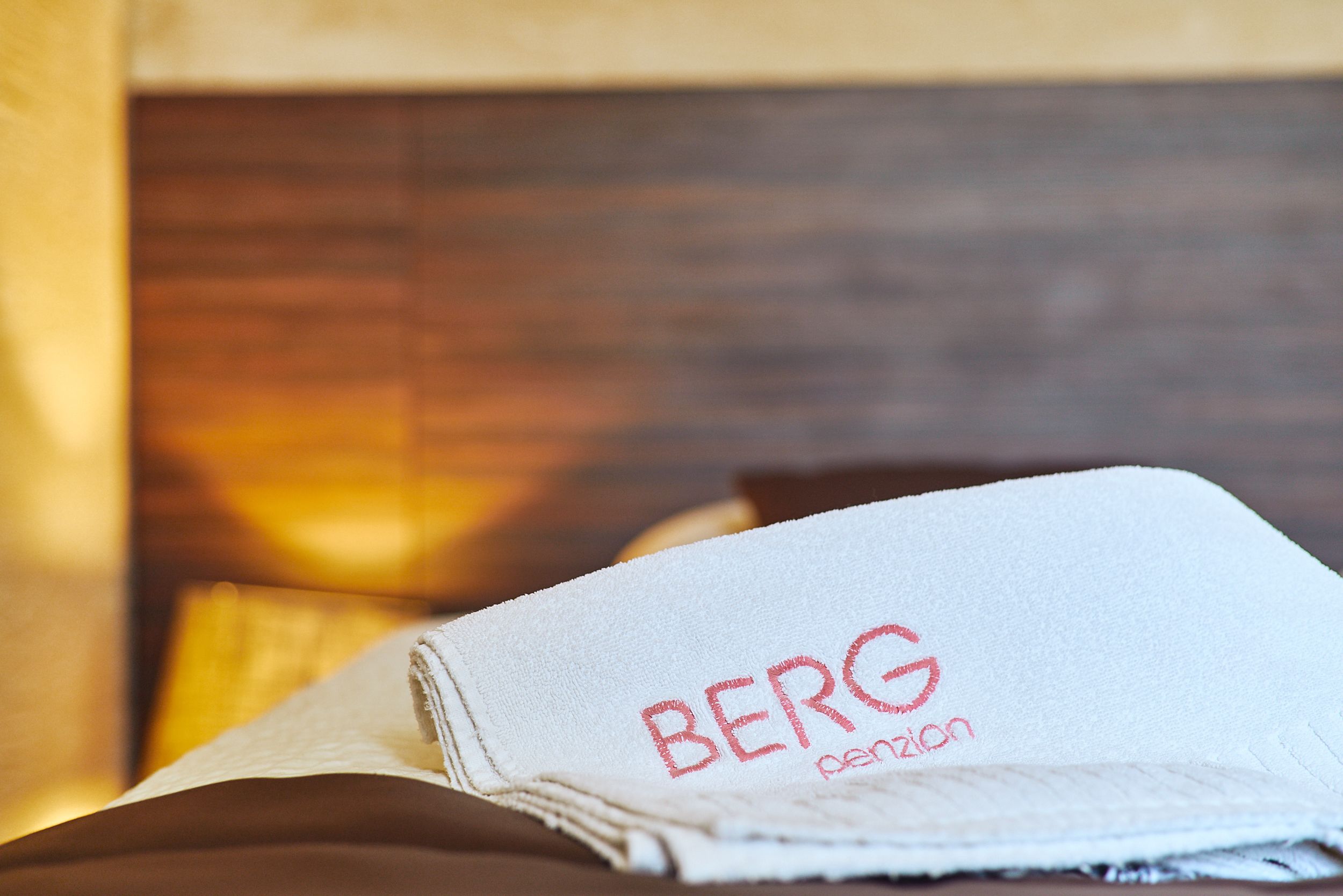 Toalha dobrada com o texto "BERG pension" sobre uma cama com cabeceira de madeira.
