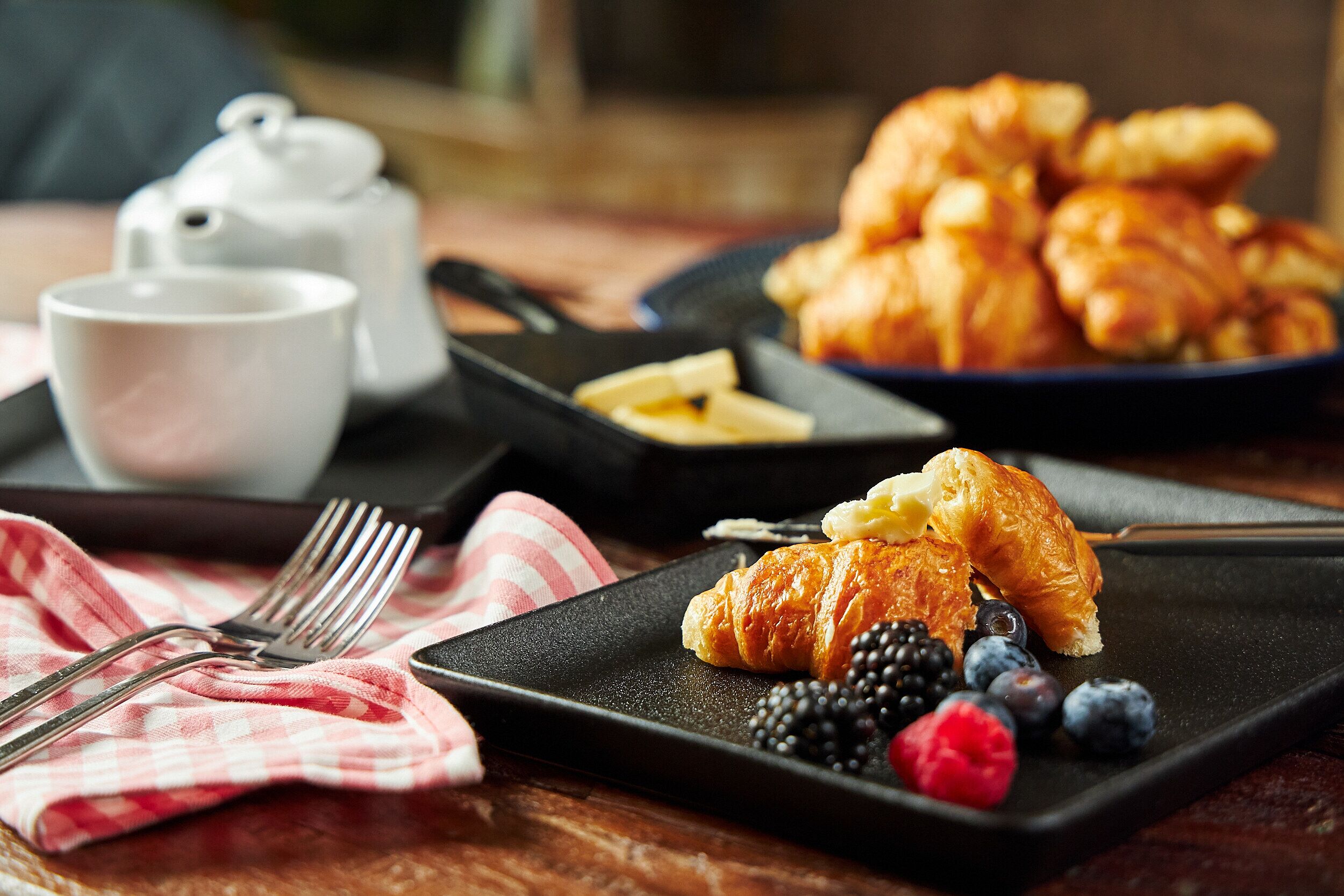 Un desayuno con croissants, arándanos, frambuesas y una tetera sobre una mesa de madera.