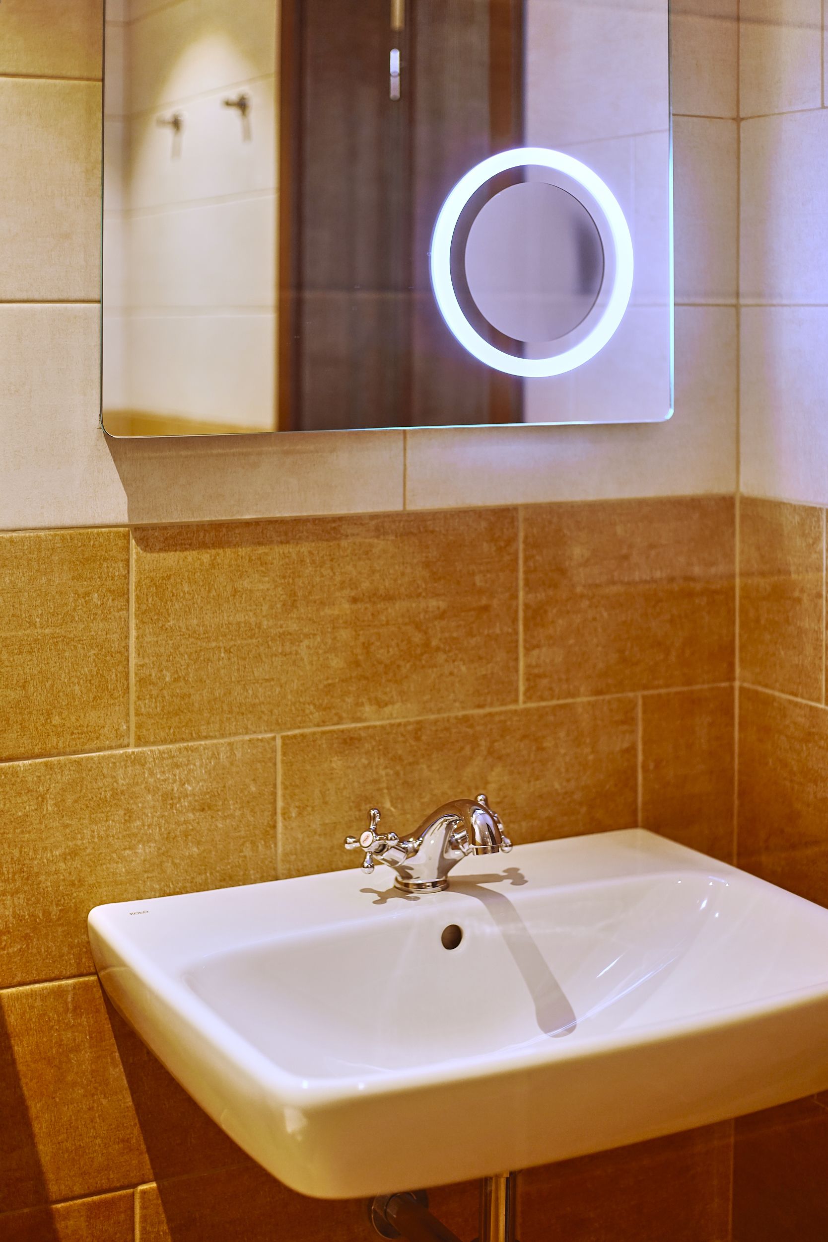 A bathroom with a white sink, a mirror with a circular light, and beige tiled walls.