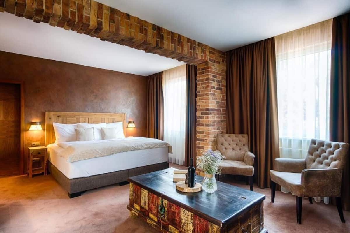 A hotel room with a large bed, two armchairs, a wooden coffee table, and a brick accent wall.