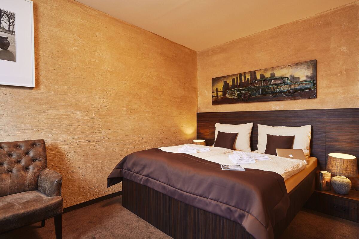 A hotel room with a bed, a chair, a painting, and a nightstand with a lamp.