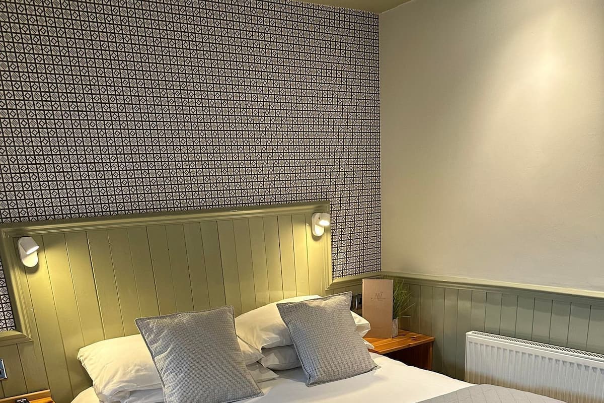 A neatly made bed with a quilted grey blanket, a headboard with wall-mounted lights, and a patterned wall above the bed.