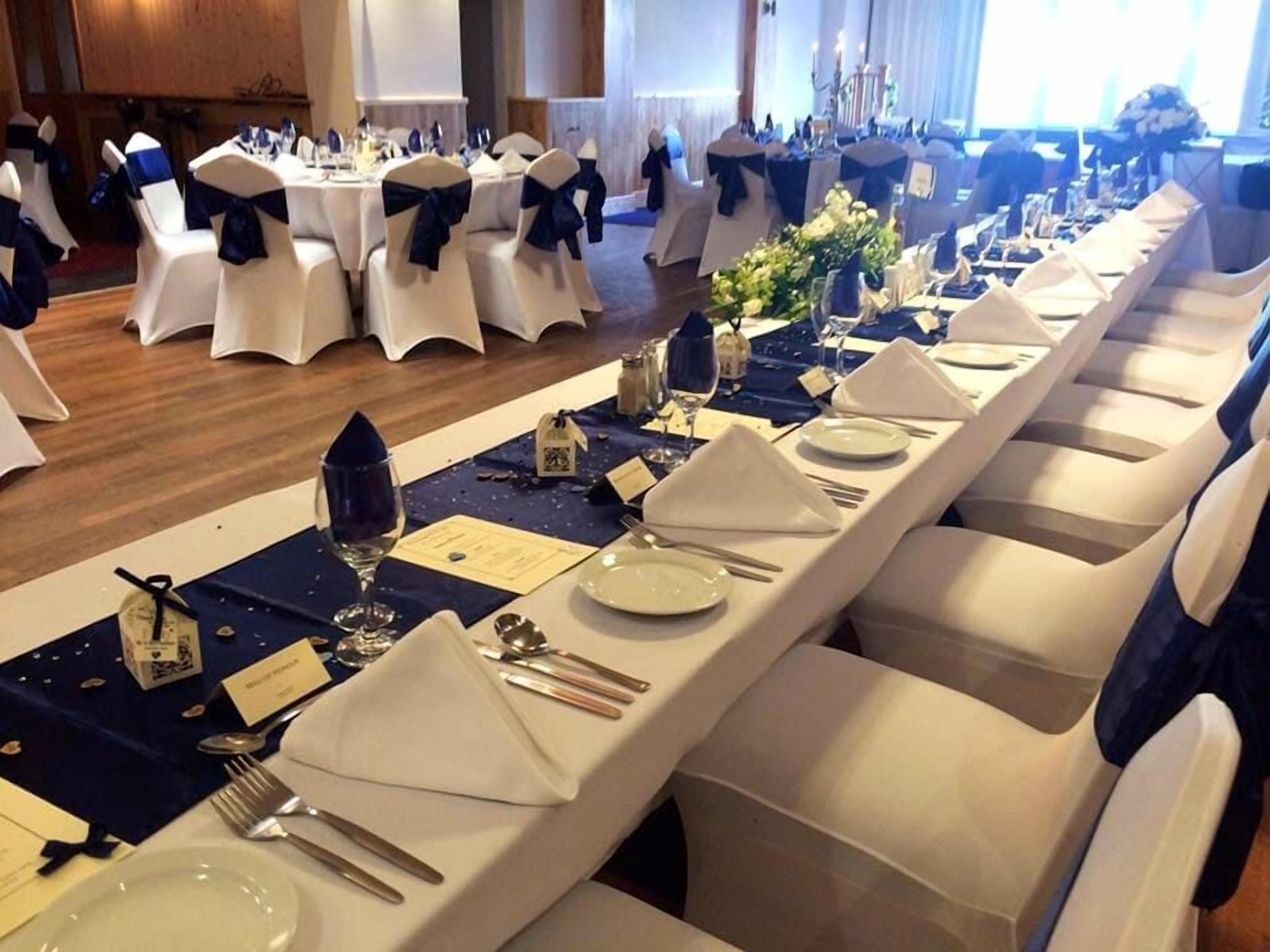 A banquet hall with long tables set for a formal event, featuring white chairs with blue sashes, neatly arranged table settings, and a chandelier in the background.