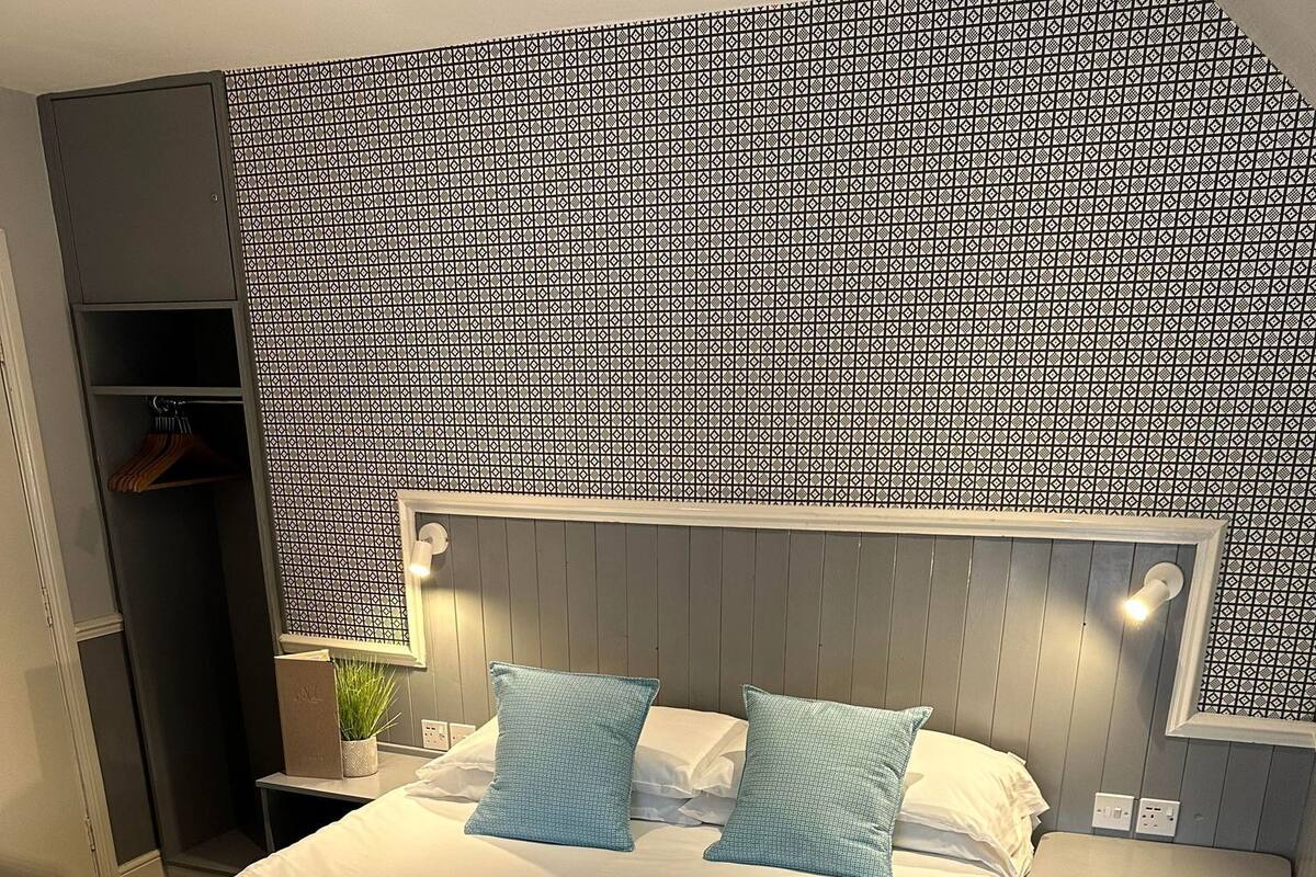 A neatly made bed with a quilted blue bedspread, two pillows, and two folded towels. There is a nightstand with a lamp and a small plant beside the bed. The room has a patterned wall and a wardrobe in the corner.