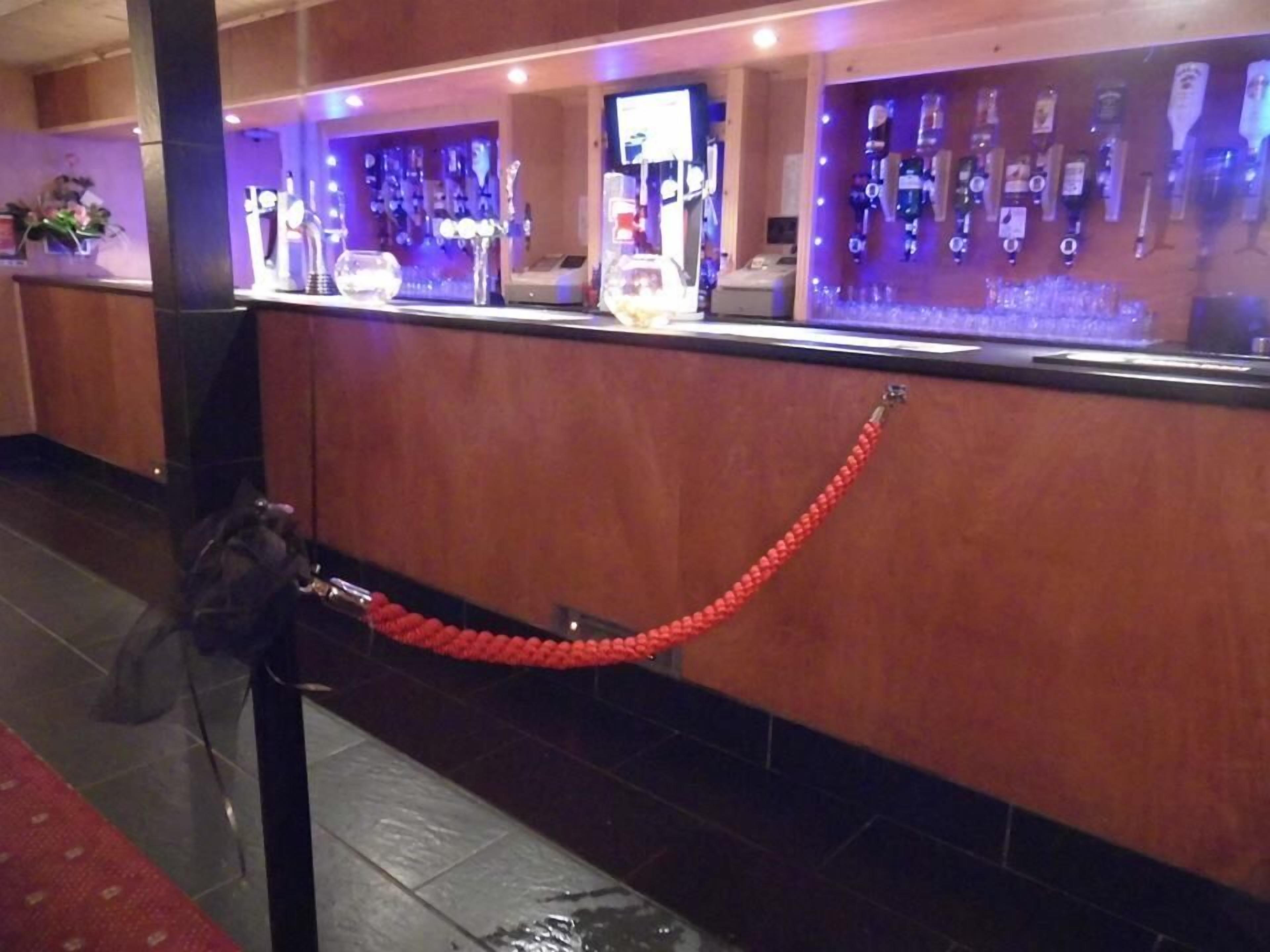 Un espace d’accueil d’hôtel avec un comptoir où sont exposées différentes bouteilles d’alcool et une barrière en corde rouge.