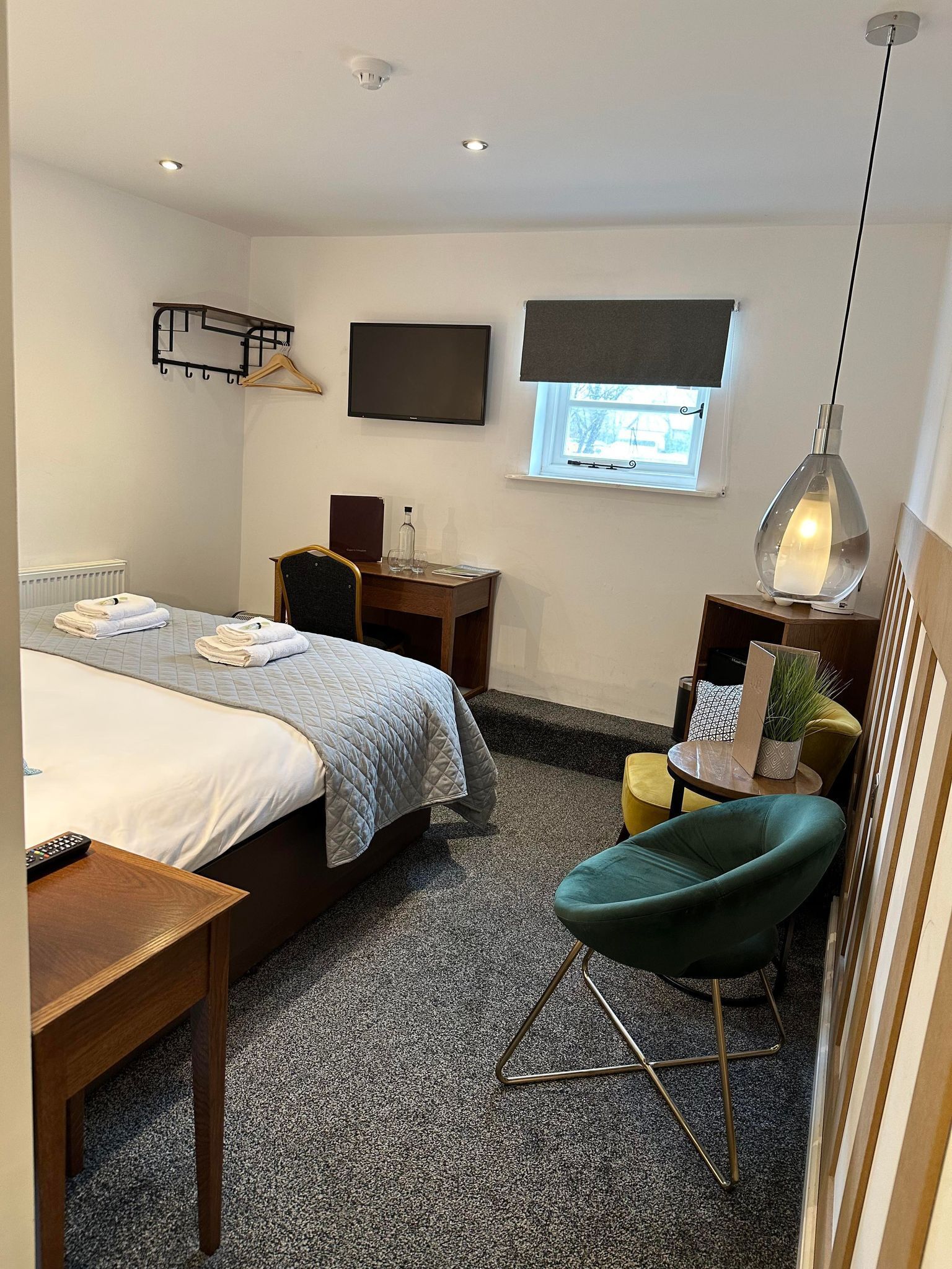 A hotel room with a bed, a desk, a chair, a TV, a window, and a hanging light fixture.
