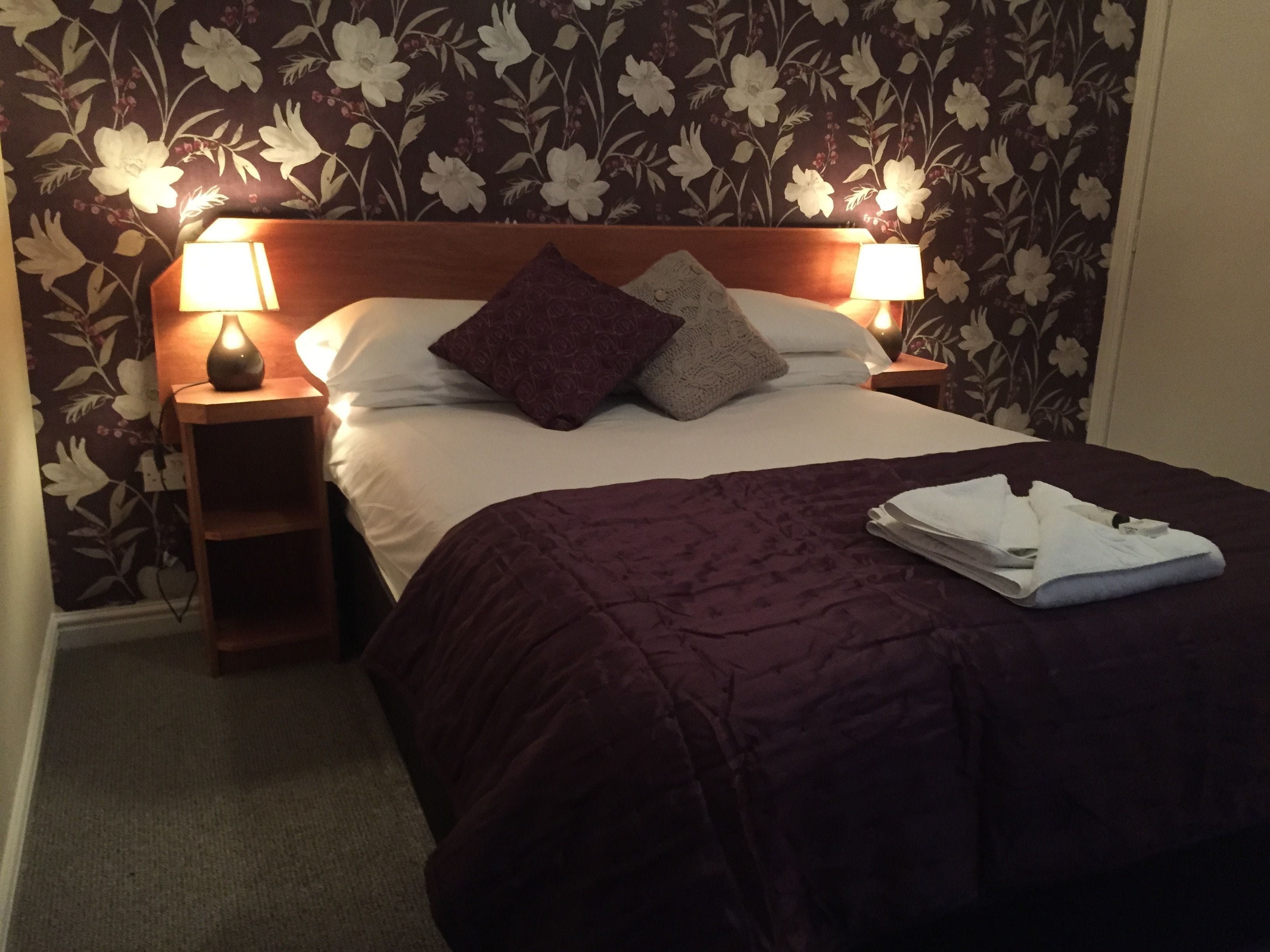 A neatly made bed with a purple bedspread and two pillows, flanked by two bedside lamps on wooden nightstands.