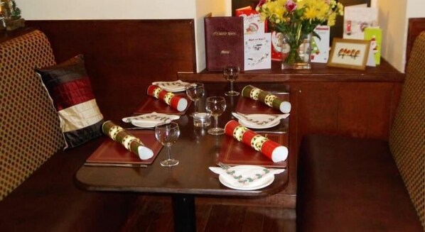 Mesa de comedor dispuesta con platos, vasos y botellas envueltas, rodeada de asientos acolchados.
