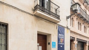 Un immeuble à la façade beige, avec une porte en bois et un balcon doté de balustrades noires. Une banderole sur l’immeuble porte l’inscription : « À LA DÉCOUVERTE DES AUBERGES DE JEUNESSE ».