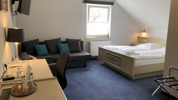 A hotel room with a bed, a desk, a chair, a sofa, a TV, a window, and a radiator.