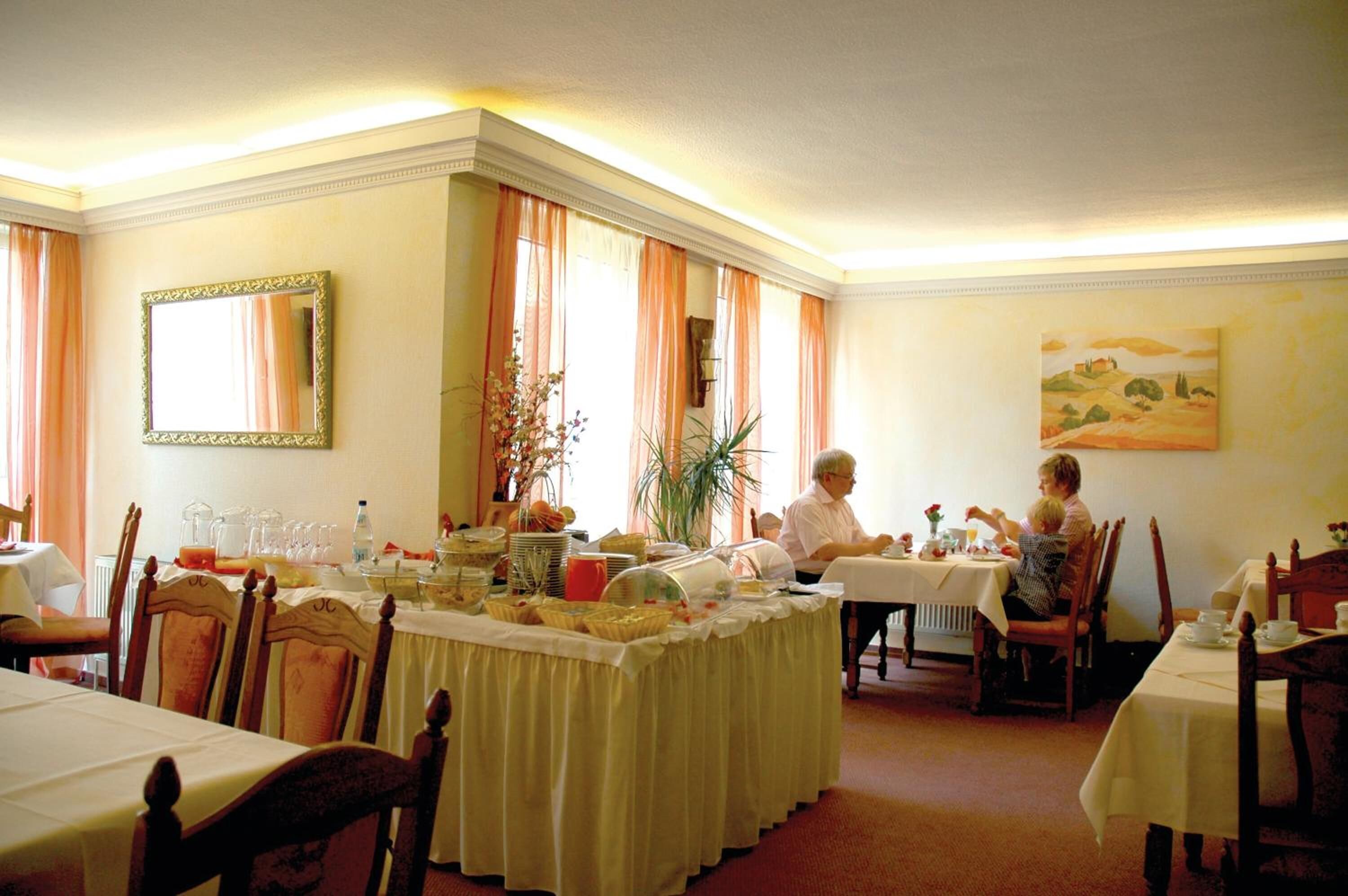 Een eethoek met gedekte tafels, een grote spiegel aan de muur en een schilderij boven de tafels.