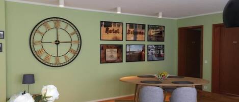 A living room with a round wall clock, a dining table with four chairs, and a sofa with a cushion.