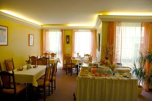 Un comedor con varias mesas dispuestas para el desayuno, mostrando diversos alimentos y una planta en maceta.