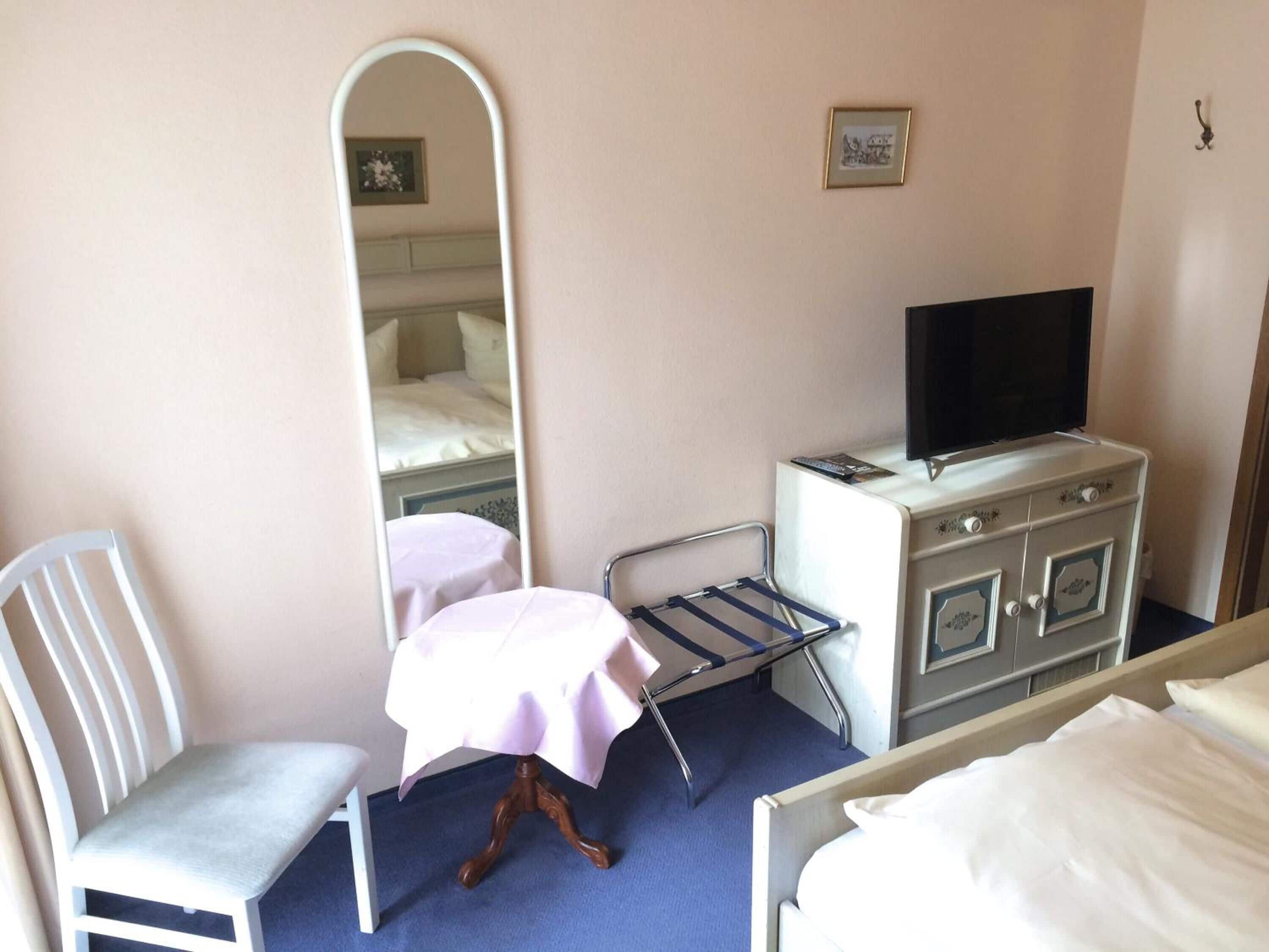 A hotel room with a bed, a chair, a small table with a tablecloth, a dresser with a television on top, and a mirror on the wall.