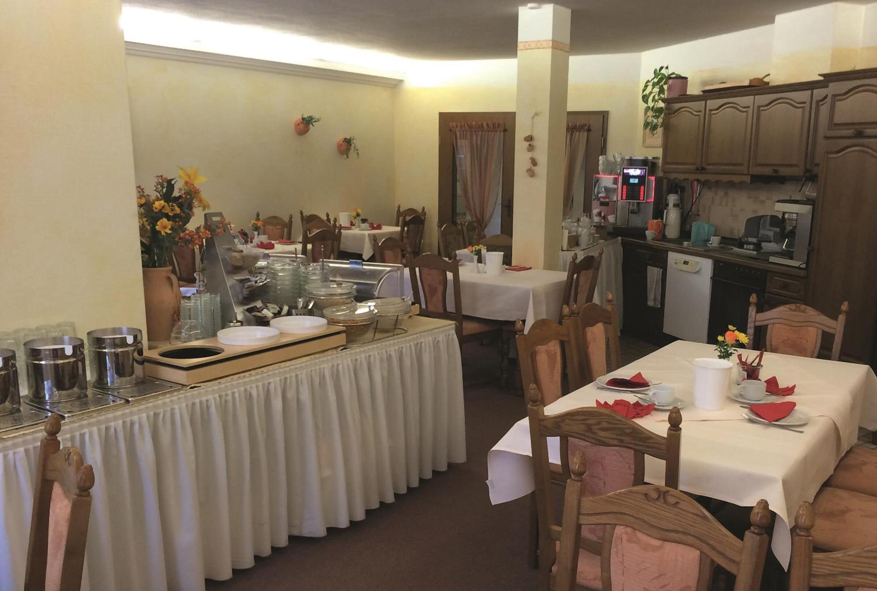 Un comedor con varias mesas dispuestas para comidas, una barra de buffet con diversos platos, y una zona de cocina con electrodomésticos y armarios.